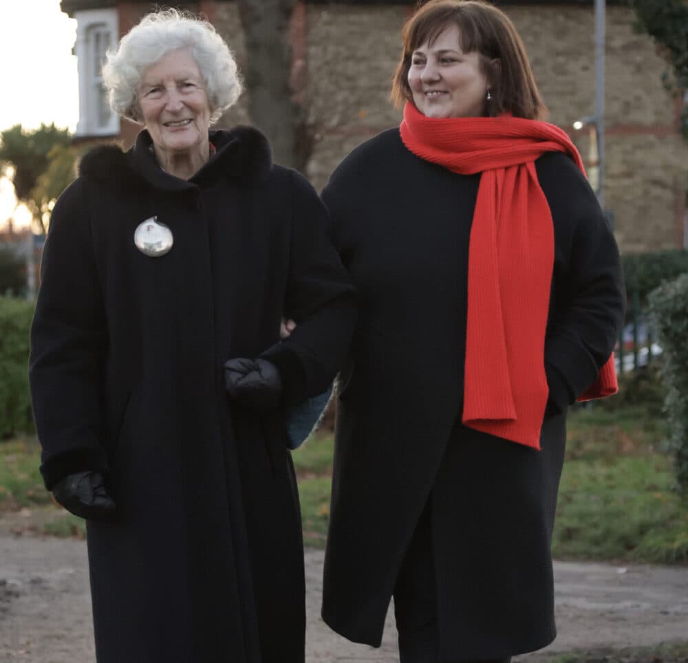 Two women walk arm in arm outdoors; one wears a red scarf and the other has white curly hair and a black coat. - Home Instead