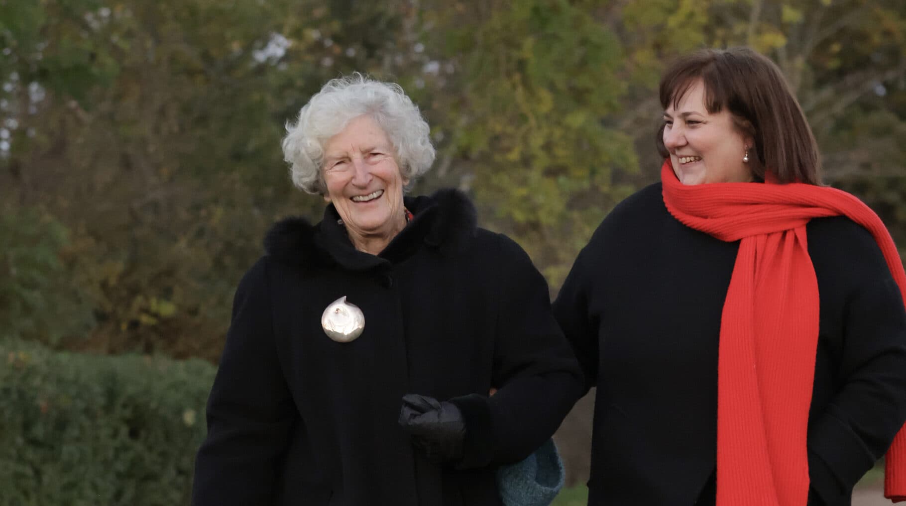 Two women in black coats, one with a red scarf, smile and walk outdoors on a fall day. - Home Instead