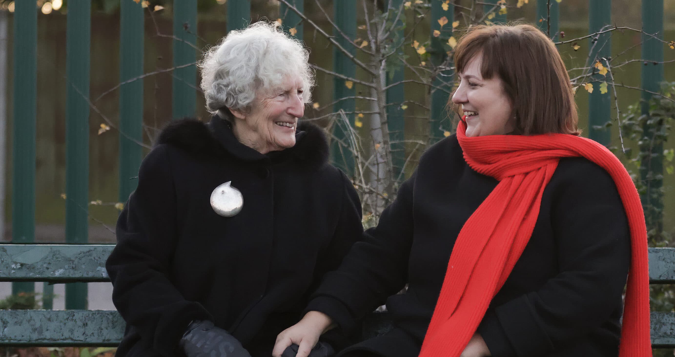 Two women in dark coats sit on a bench, smiling at each other; one wears a bright red scarf. - Home Instead