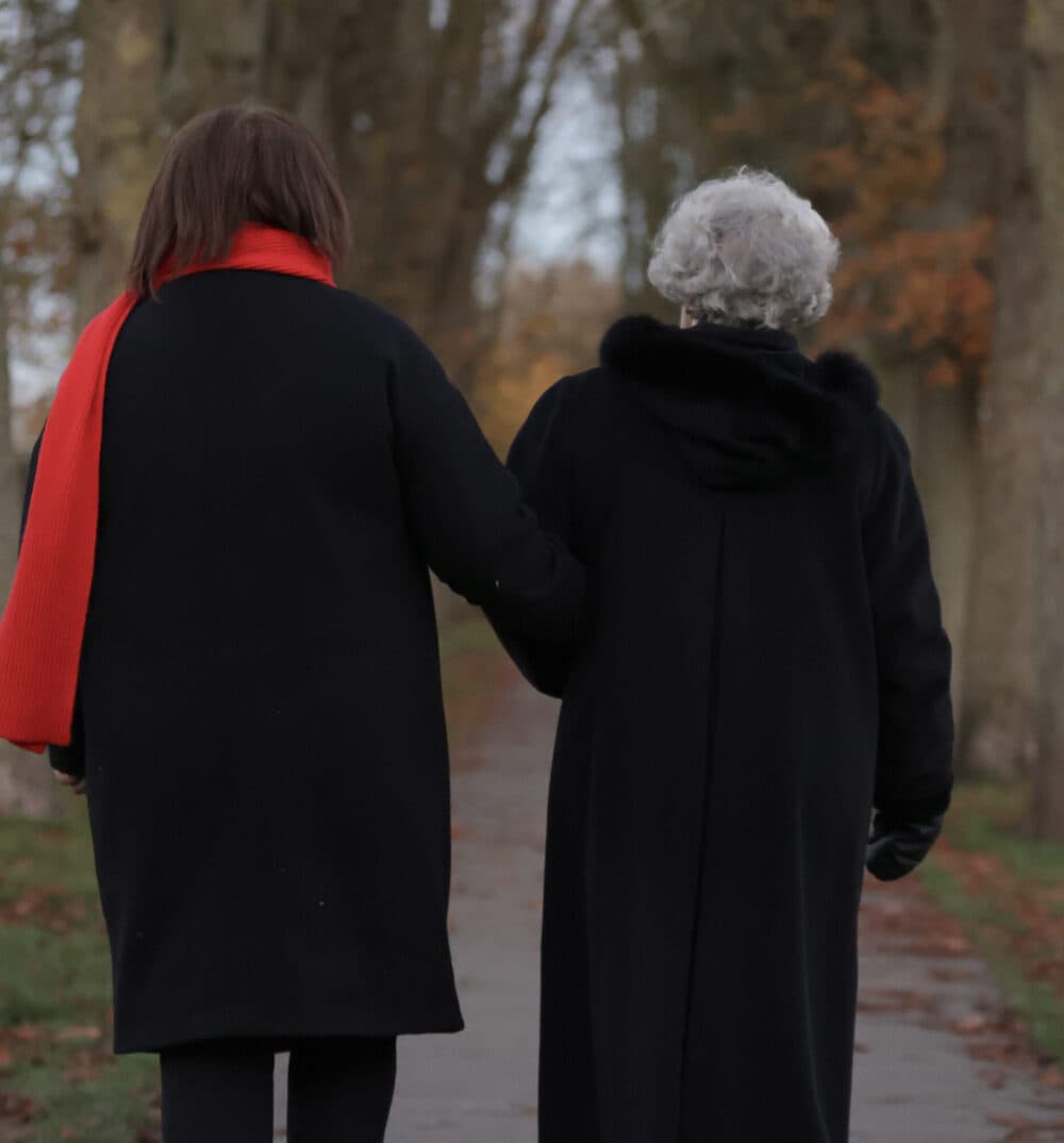 Two people in dark coats, one wearing a red scarf, walk arm in arm down a tree-lined path. - Home Instead