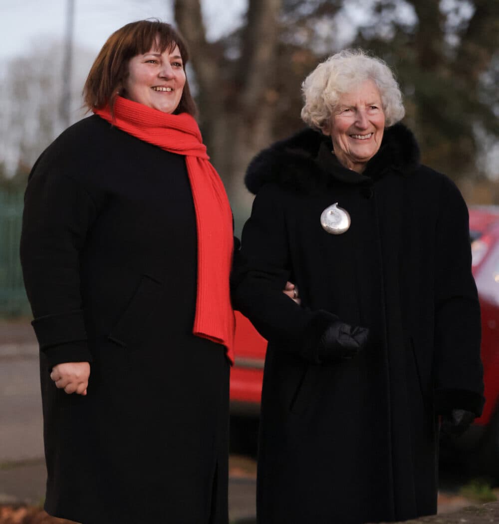 Two women in black coats, one with a red scarf, smiling and standing arm in arm outdoors. - Home Instead