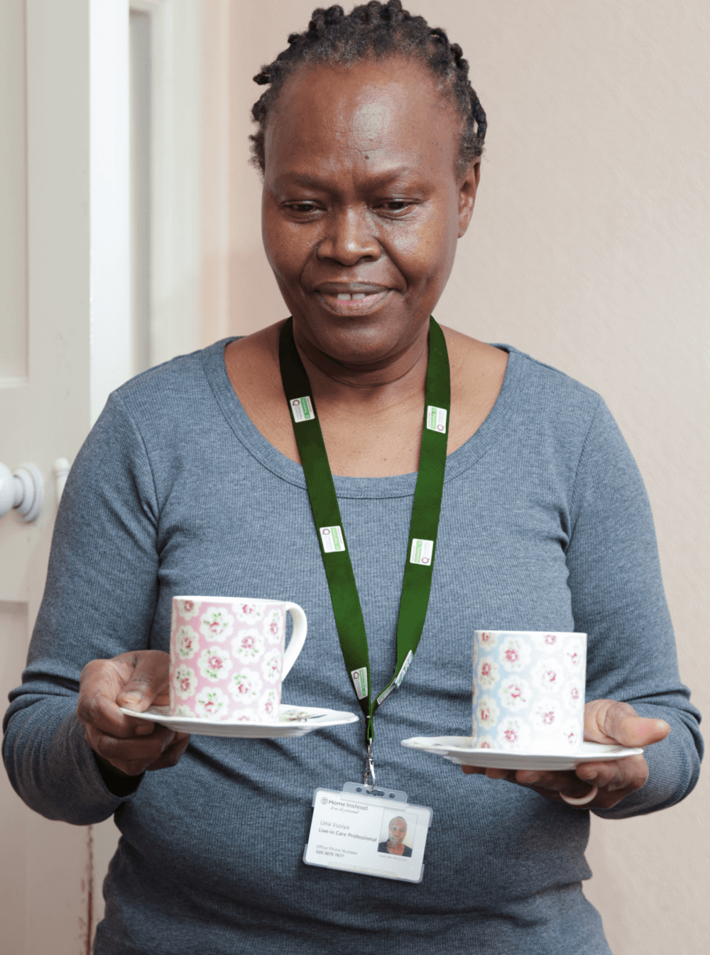 A woman holding two floral teacups on saucers, wearing a gray shirt and a green lanyard. - Home Instead