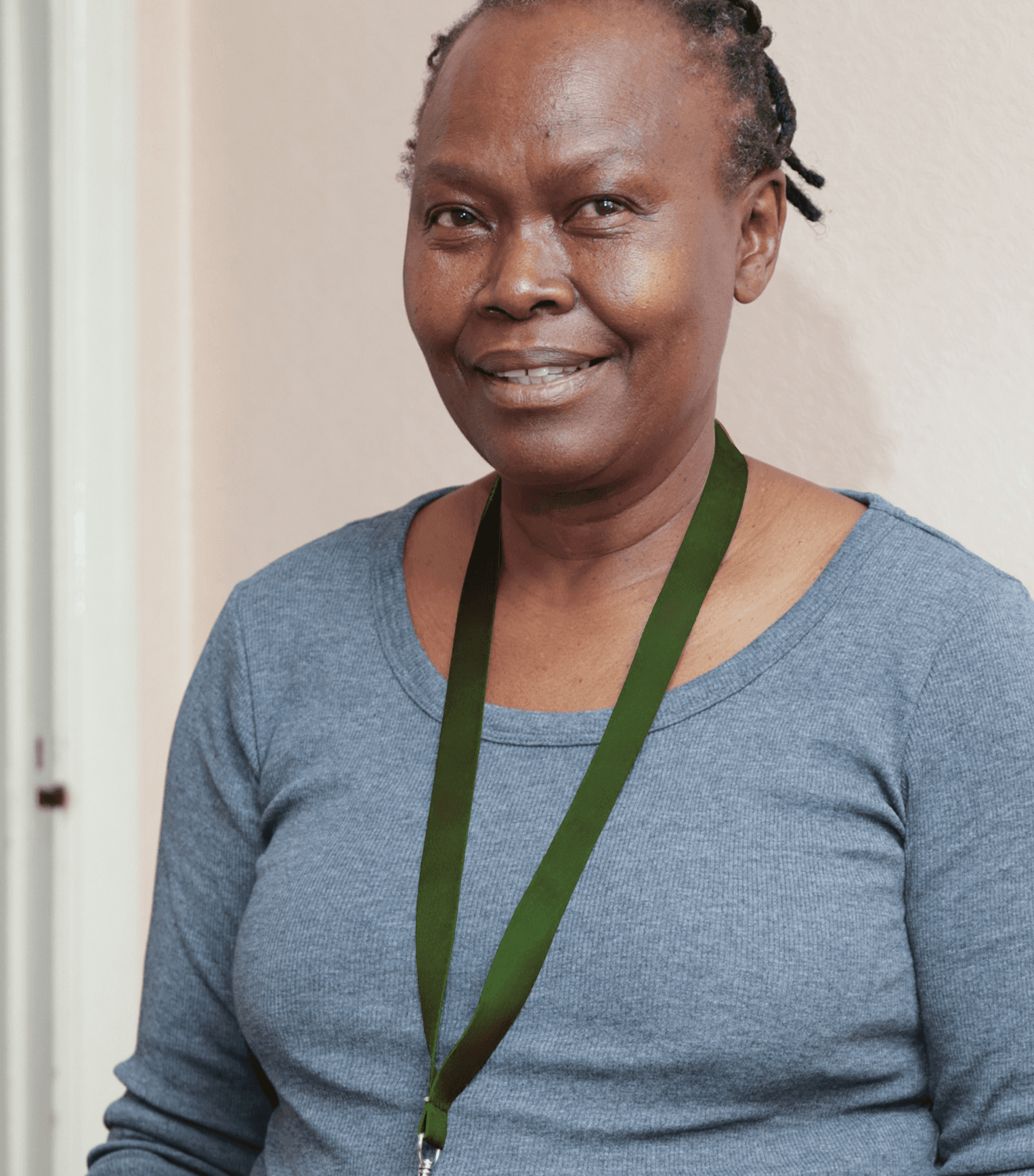 Smiling woman in a grey top wearing a green lanyard, standing indoors against a light-colored wall. - Home Instead