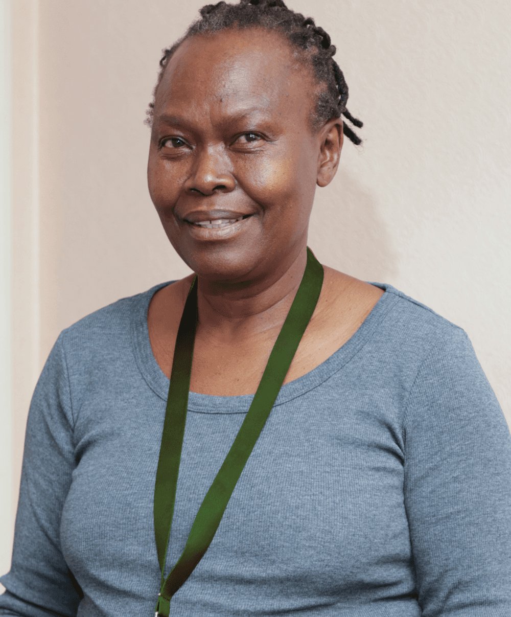 Smiling woman with short braided hair, wearing a gray top and a green lanyard, stands indoors. - Home Instead