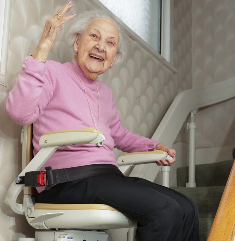Smiling elderly woman waving while seated on a stairlift indoors next to a staircase. - Home Instead