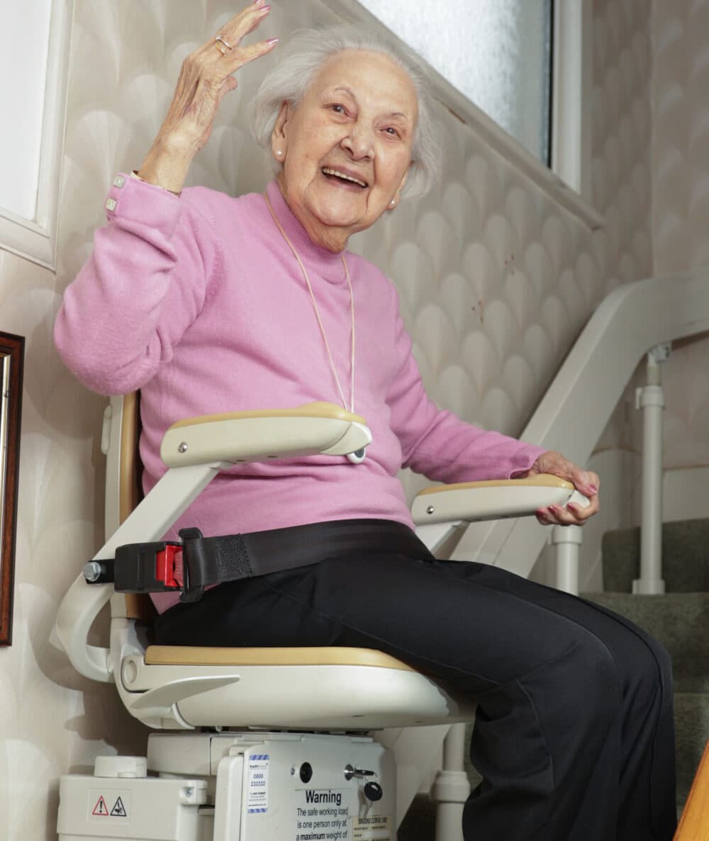 Smiling elderly woman in a pink sweater sits on a stairlift, waving at the camera inside a home. - Home Instead
