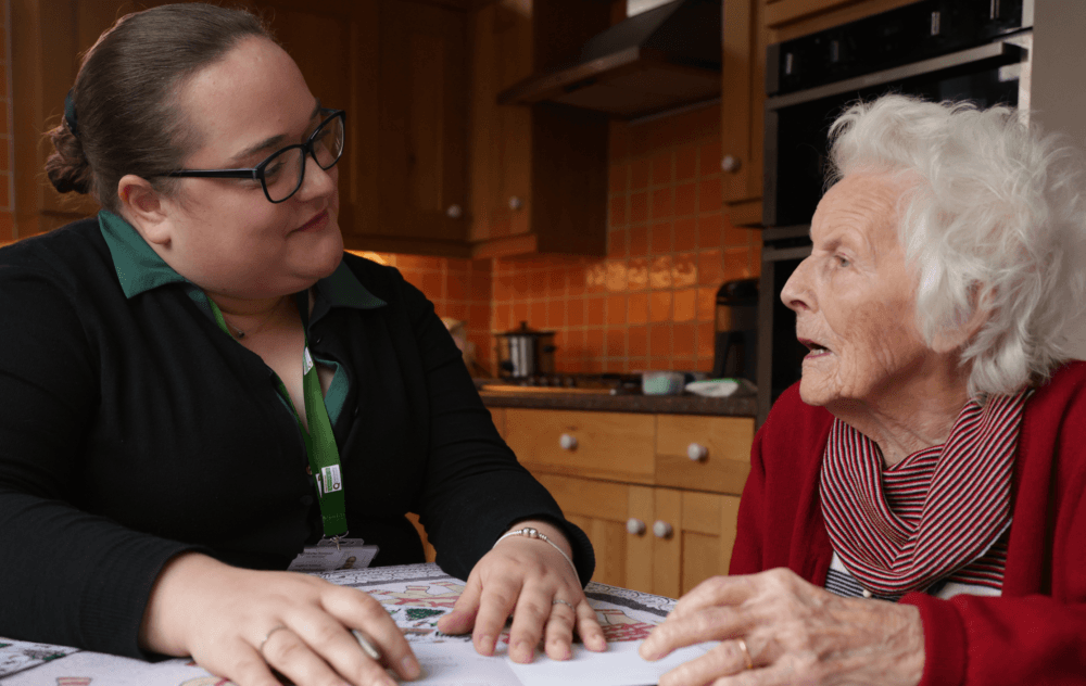 A caregiver sits at a kitchen table talking with an elderly woman in a red sweater. - Home Instead