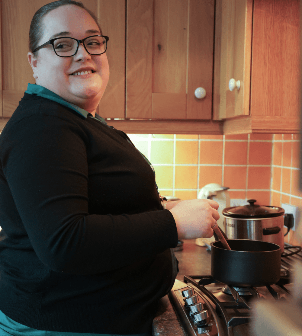 A woman smiles while stirring a pot on a stovetop in a cozy kitchen. - Home Instead