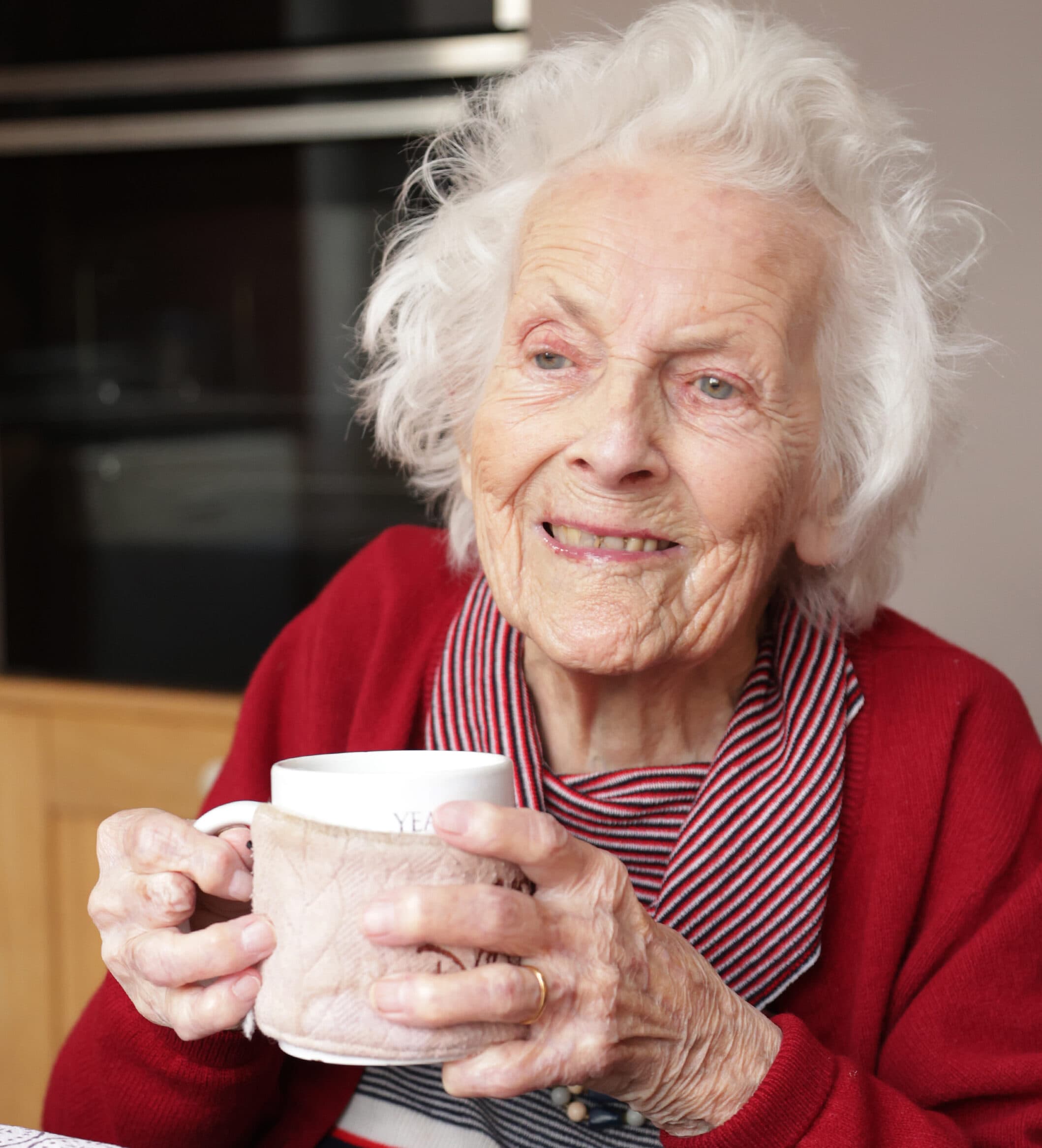 Smiling elderly woman with white hair holding a mug, wearing a red sweater and striped scarf. - Home Instead
