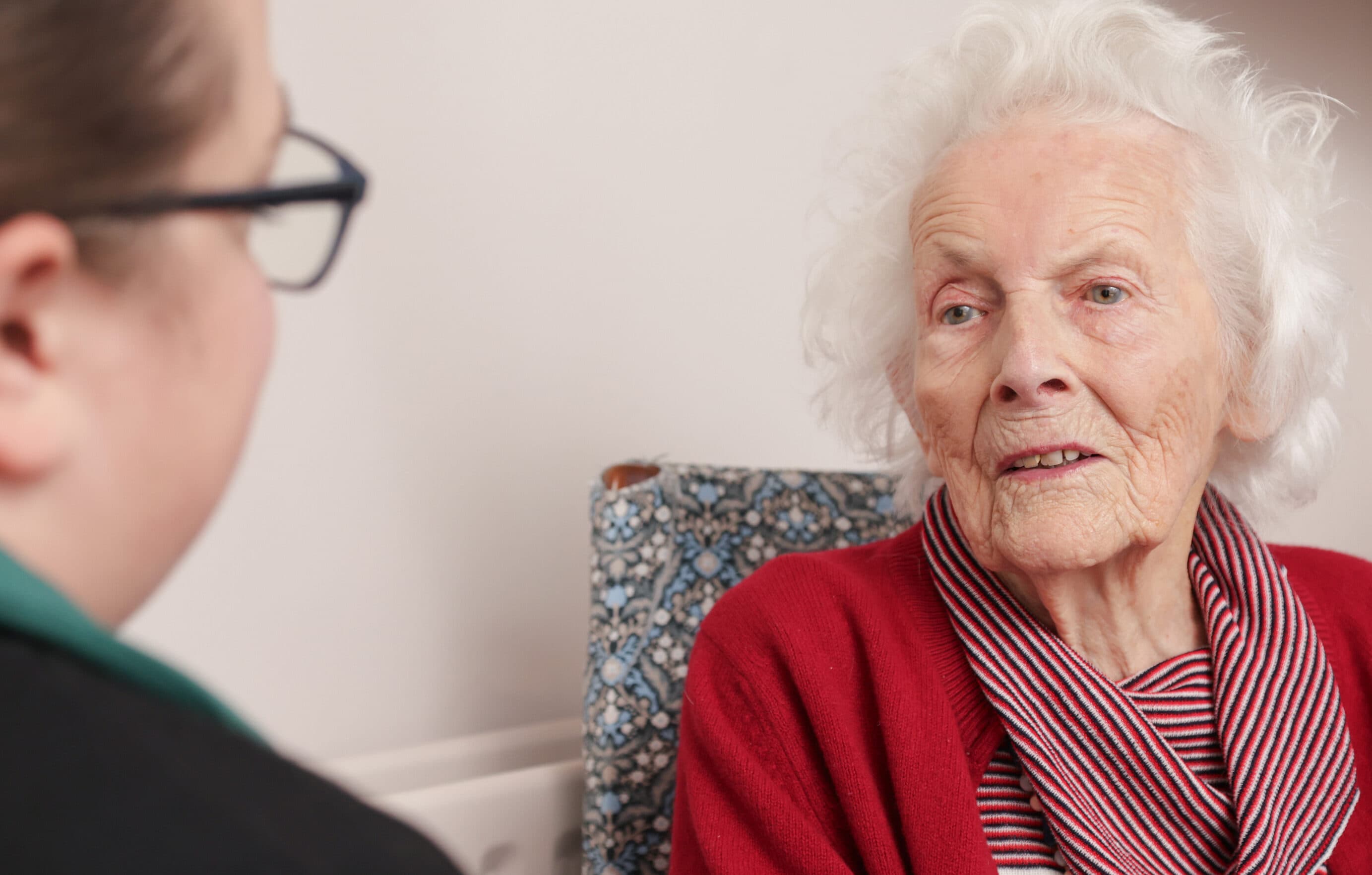 An elderly woman in a red sweater talks to a younger woman with glasses in a close indoor setting. - Home Instead