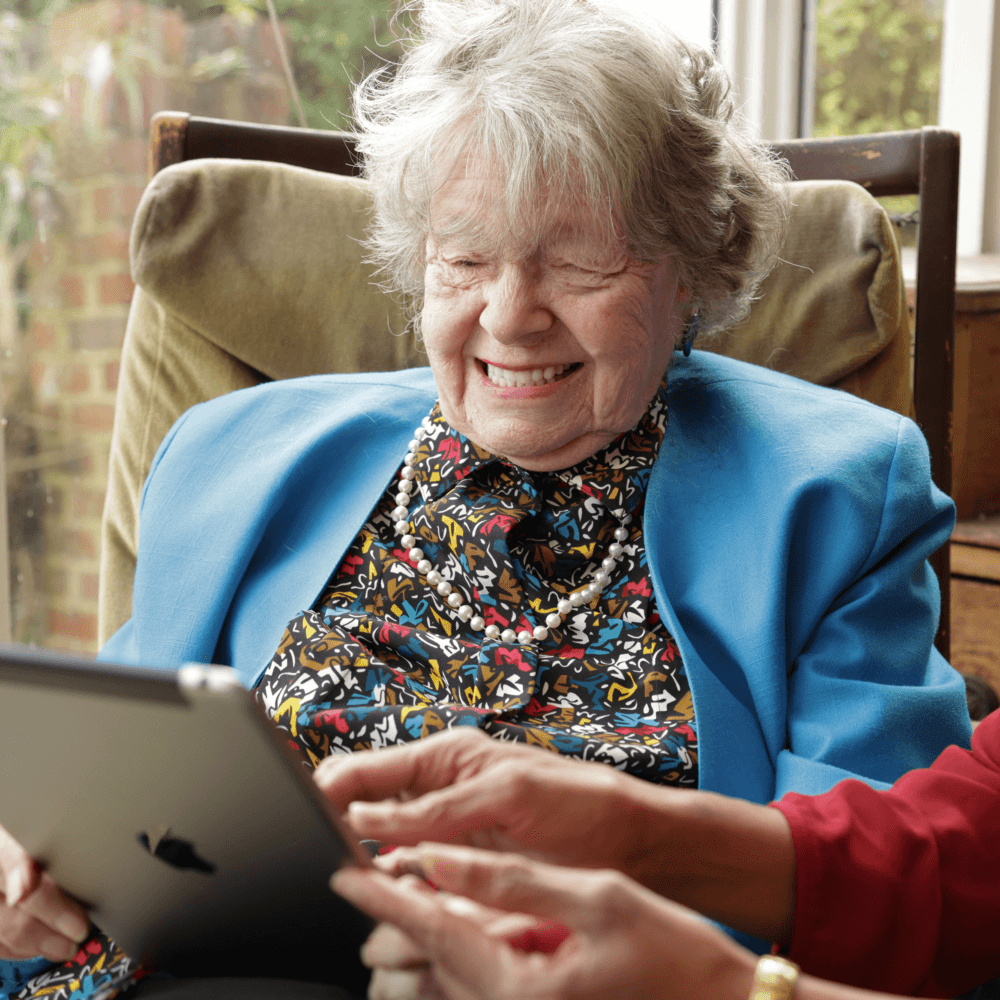 Smiling elderly woman in a blue jacket using a tablet with help from another person. - Home Instead