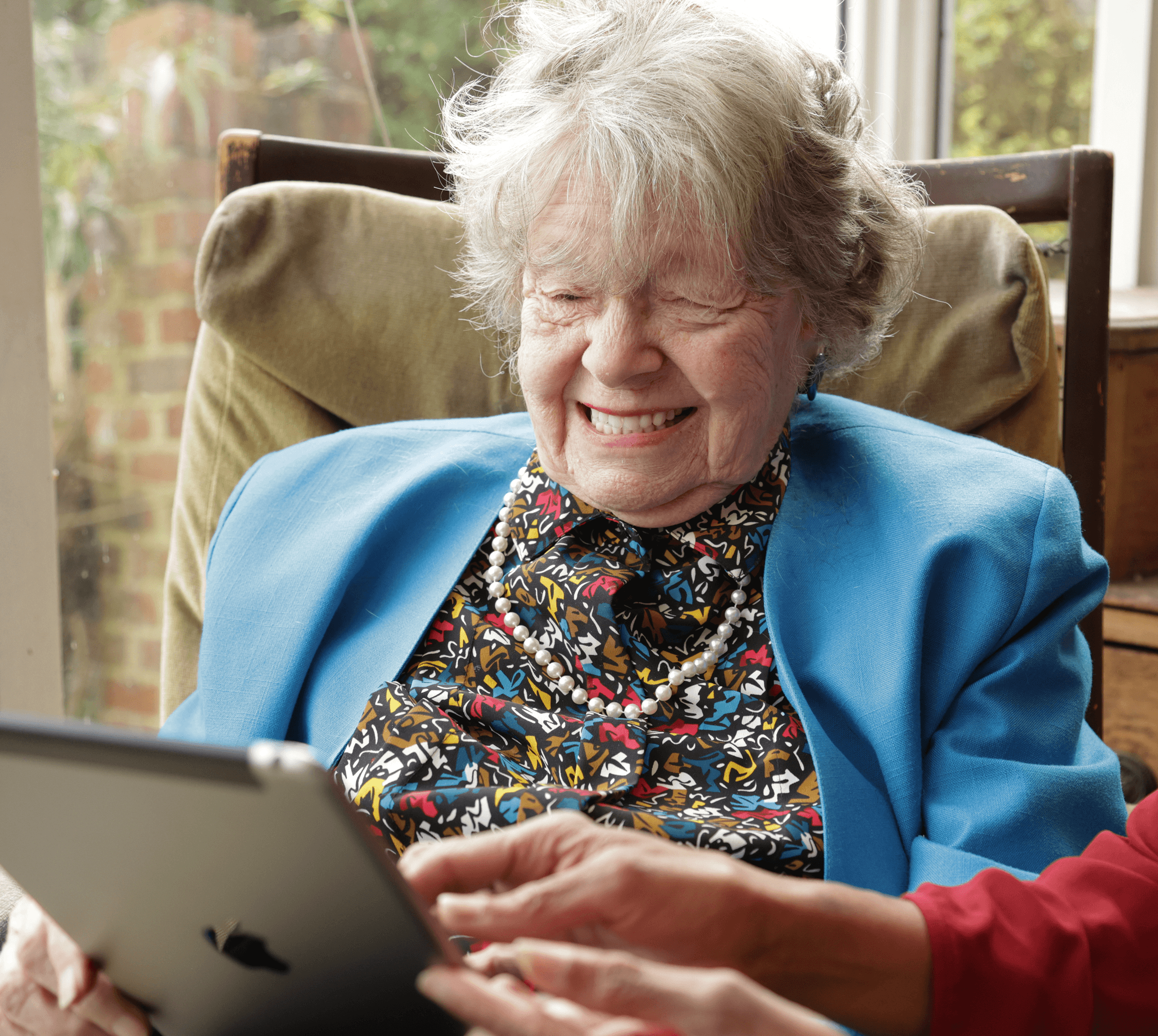 Smiling elderly woman in a blue jacket using a tablet, assisted by another person’s hands. - Home Instead