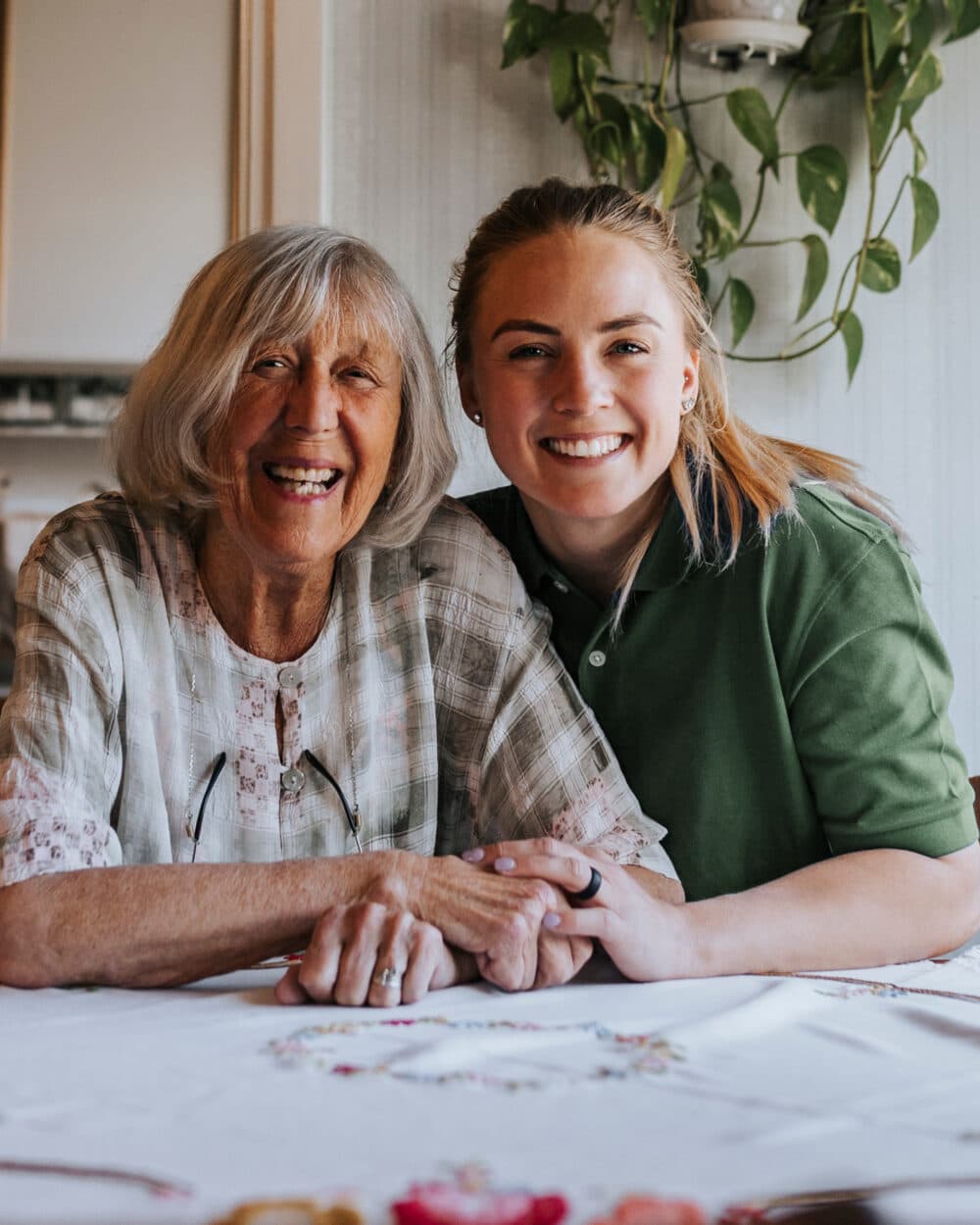 Elderly woman and young woman smiling together at a table, holding hands, indoors with a plant in the background. - Home Instead