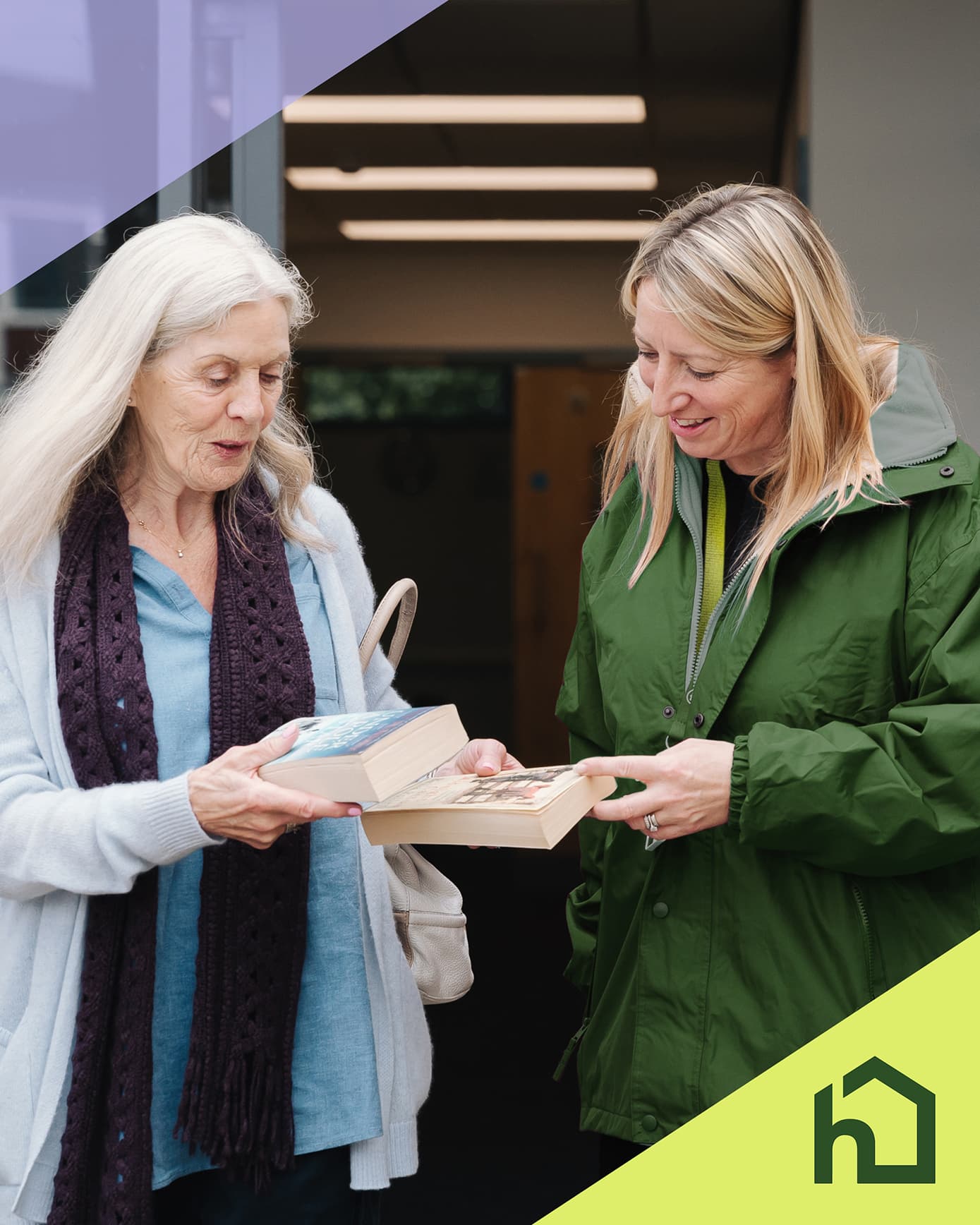 Two women standing and smiling while exchanging books indoors, with a green house icon in the corner. - Home Instead