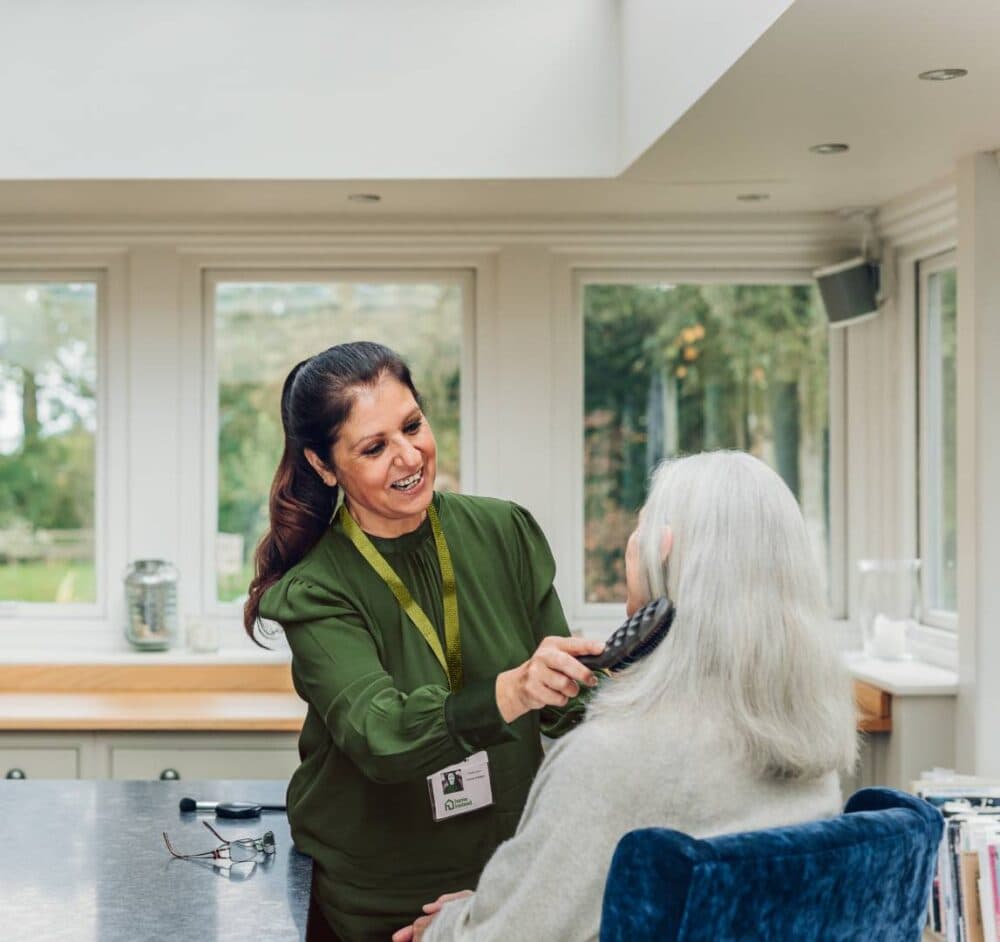 A caregiver in a green shirt brushes an older woman's gray hair in a bright room with large windows. - Home Instead
