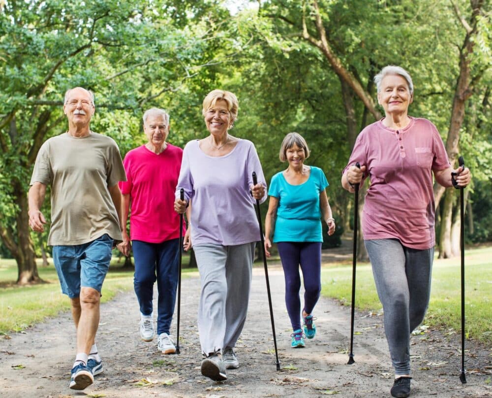 Five older adults walking with poles on a wooded path, smiling and enjoying outdoor exercise together. - Home Instead