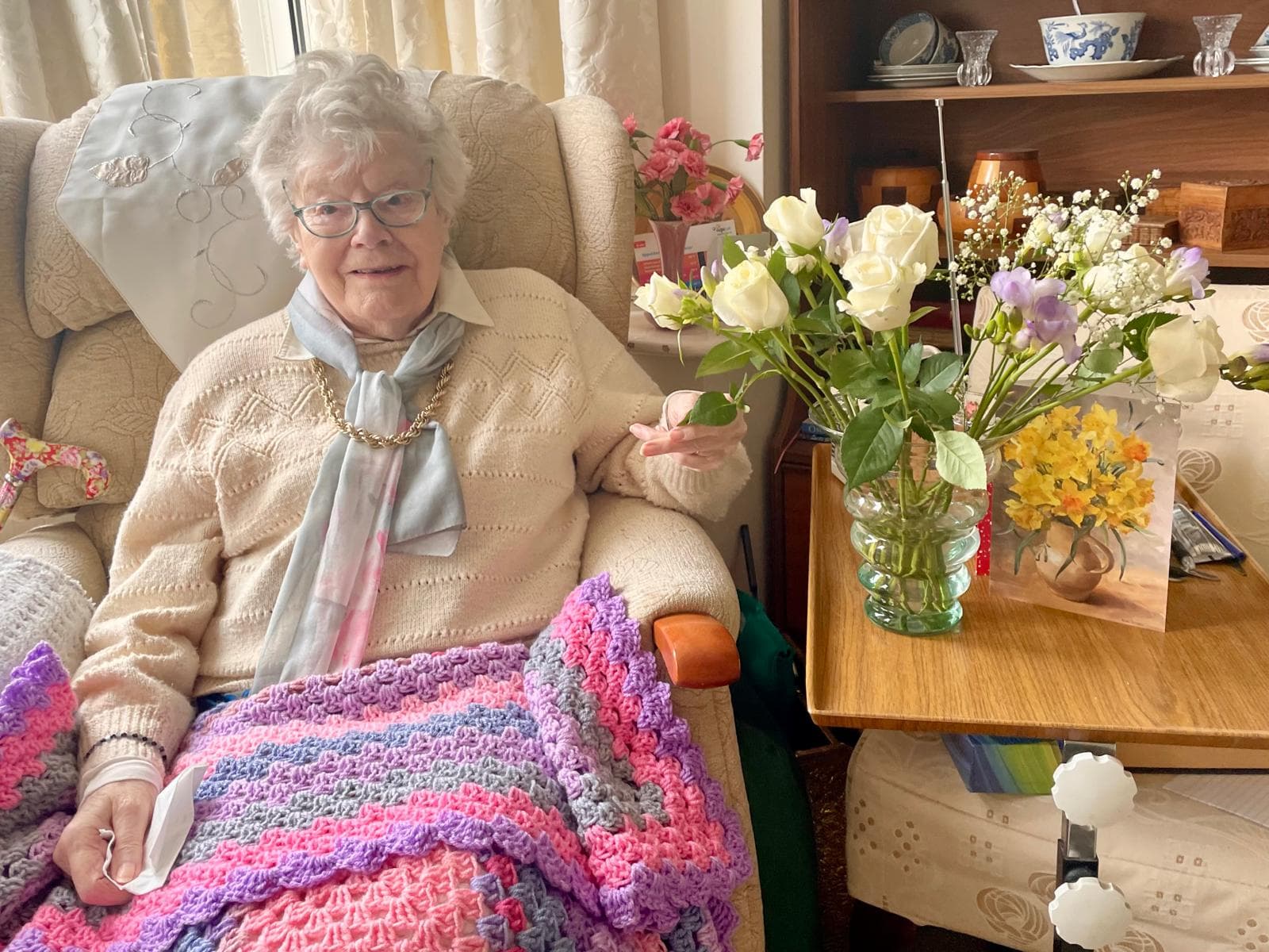 Elderly woman smiling in an armchair with a colorful blanket and a table of flowers beside her. - Home Instead