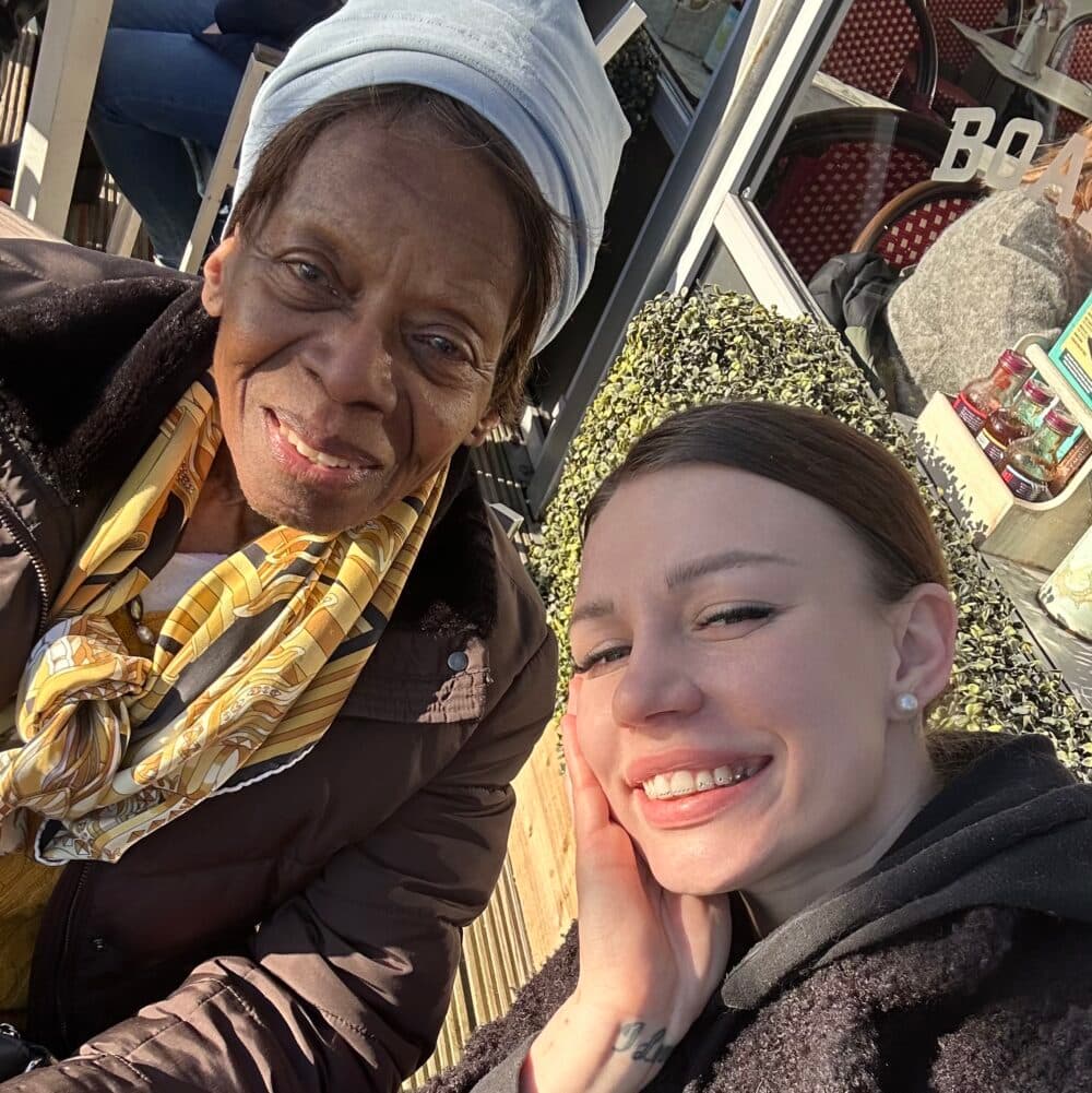 An older woman and a young woman smiling together outdoors in sunlight. - Home Instead