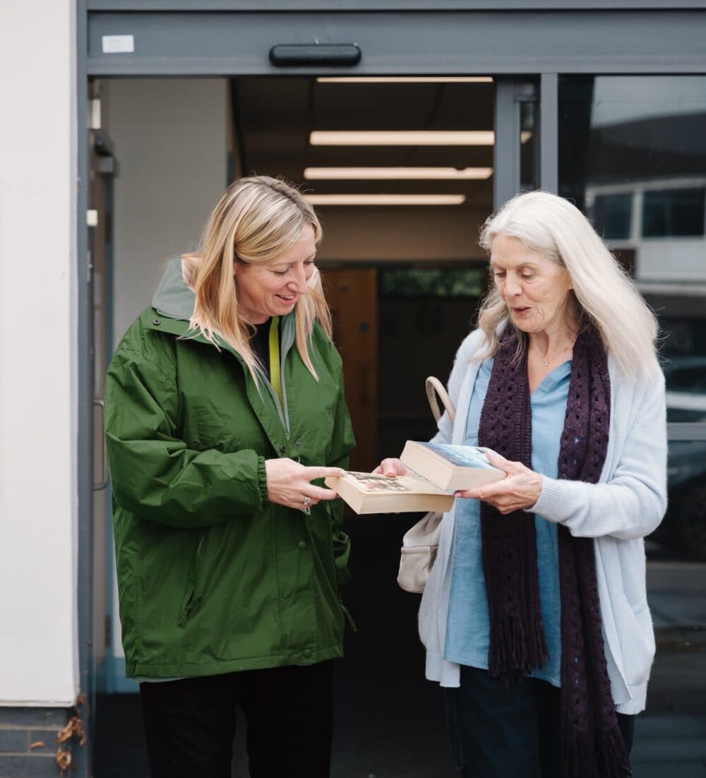 Two women standing outside a building, exchanging books and smiling at each other. - Home Instead