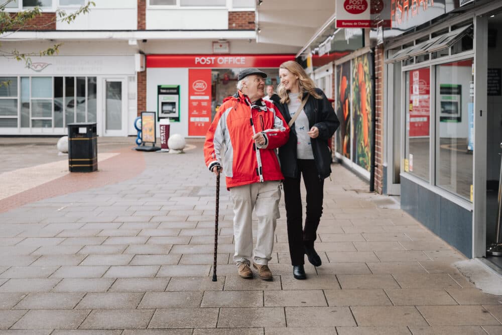 An elderly man with a cane walks and smiles with a woman on a paved sidewalk near a post office. - Home Instead