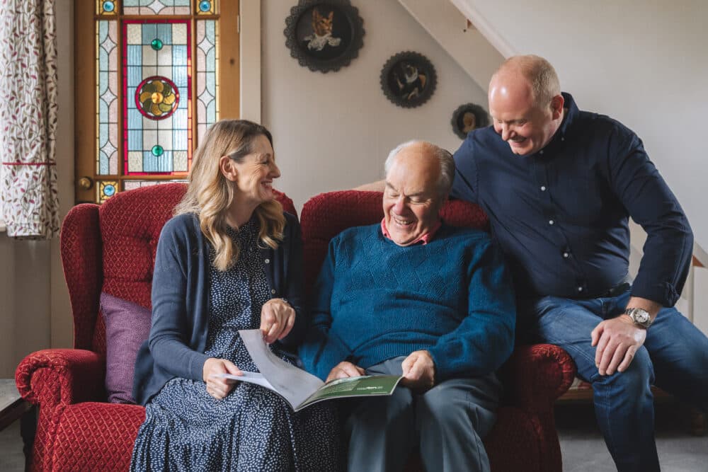 Three people sit smiling on a red sofa, looking at a booklet together in a cozy, well-lit room. - Home Instead