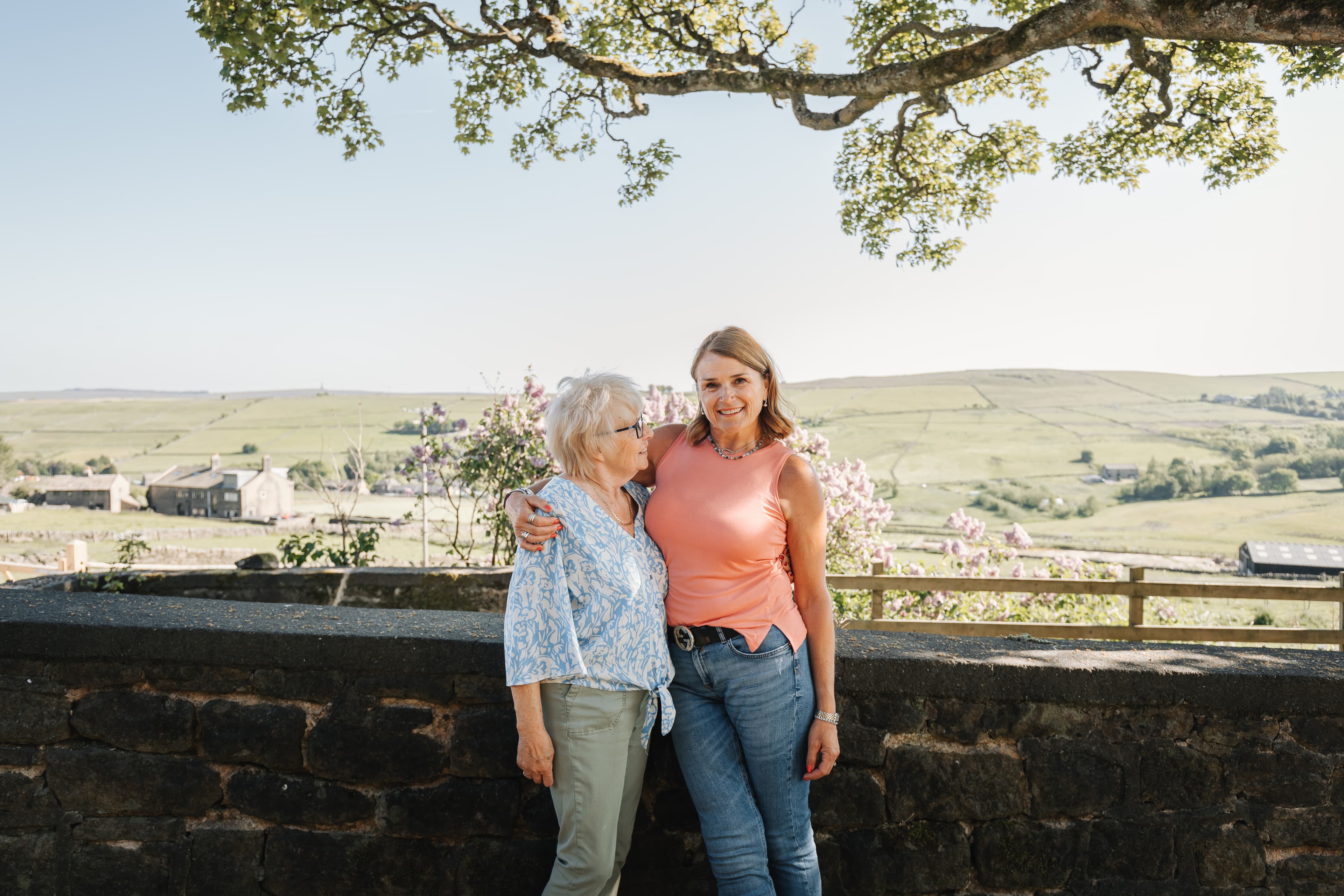 Two women standing and smiling together by a stone wall with a scenic countryside view behind them. - Home Instead