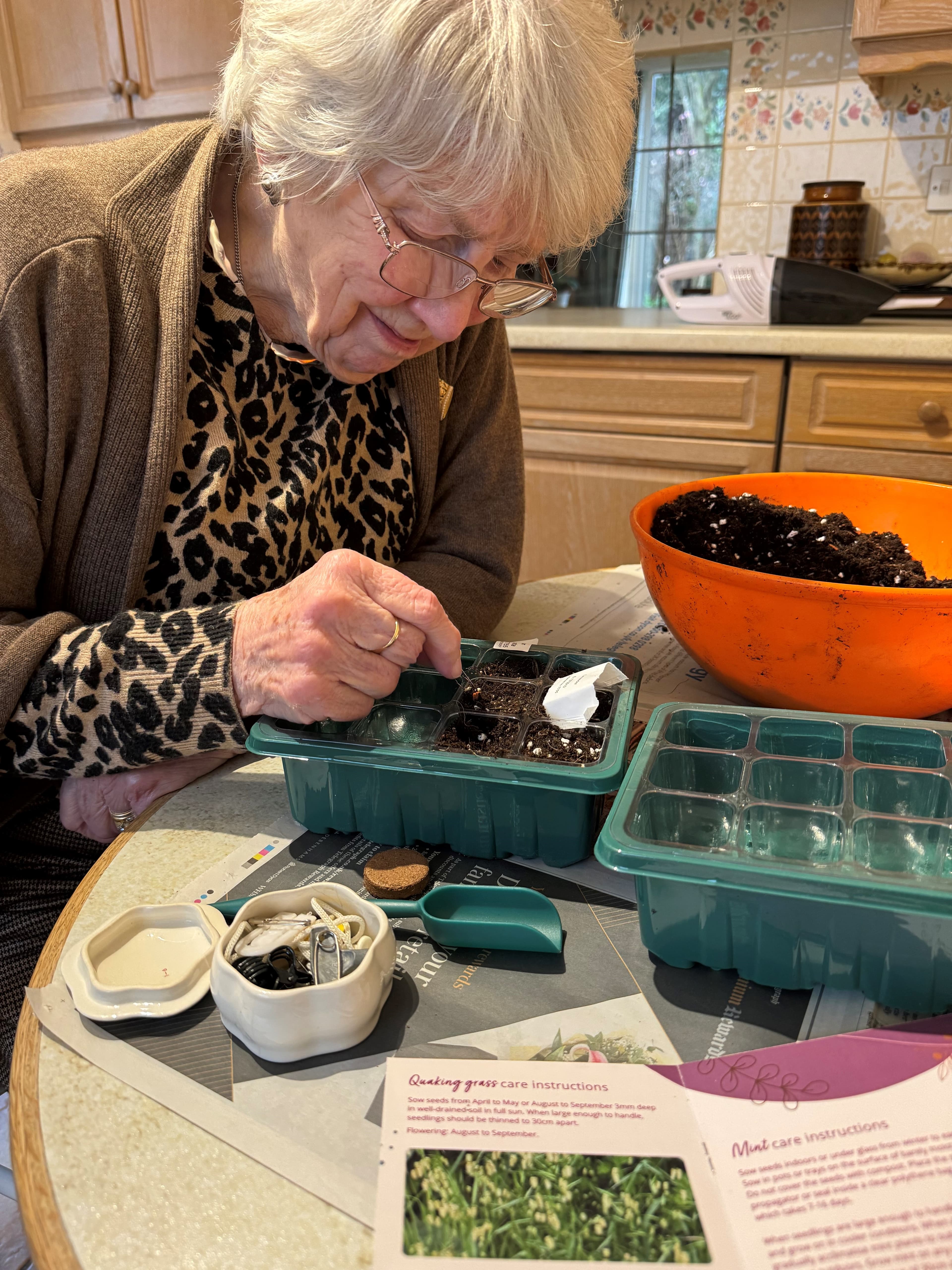Elderly woman planting seeds in seed trays at a kitchen table with soil and gardening supplies. - Home Instead