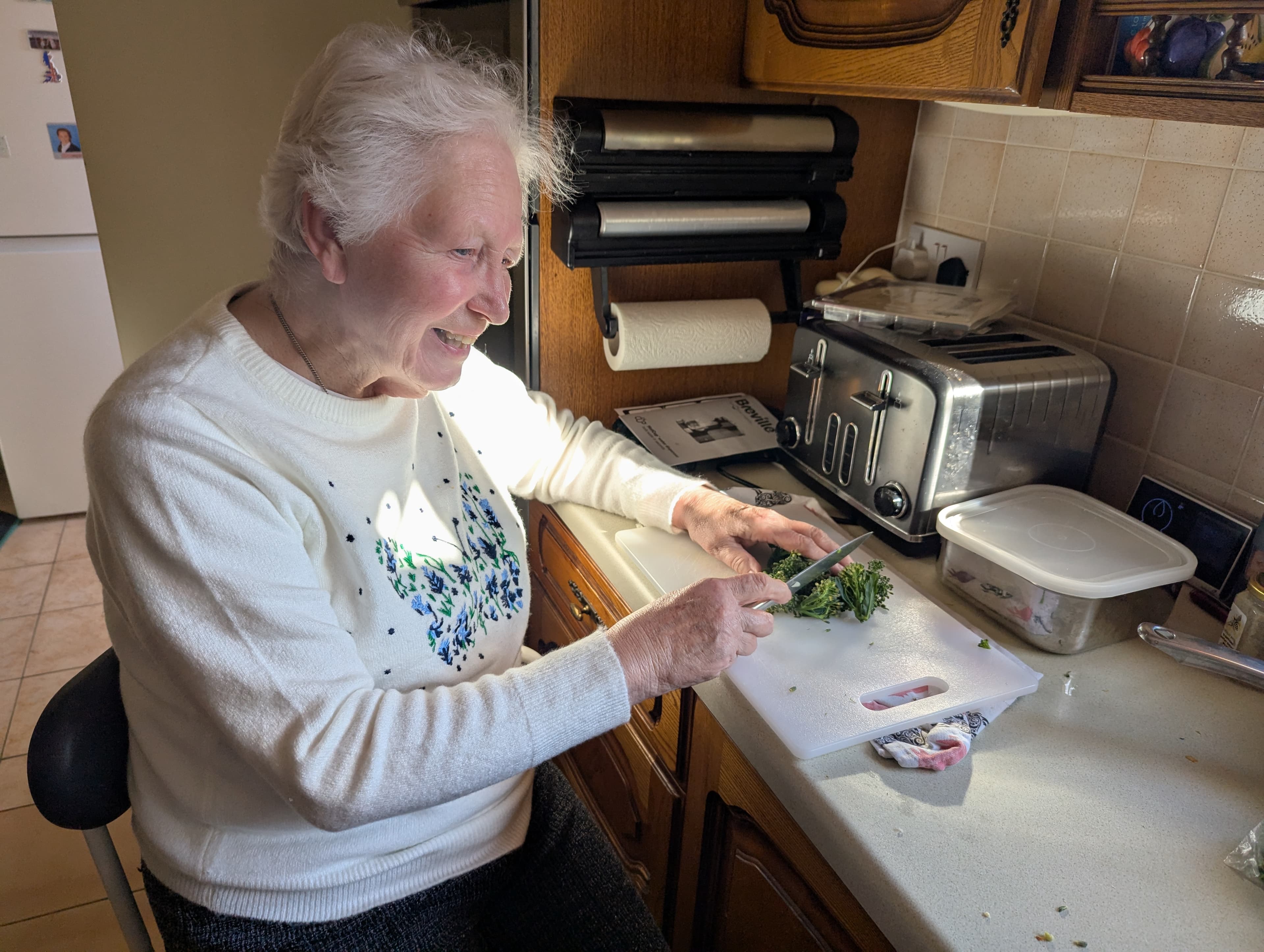 Smiling elderly woman chopping herbs on a cutting board in a sunlit kitchen. - Home Instead
