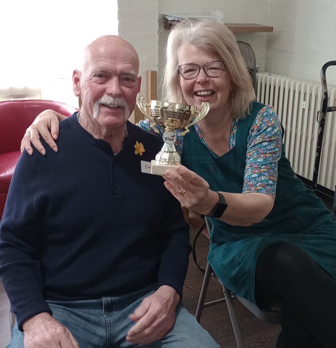 Smiling man and woman sitting, holding a gold trophy together in a bright room. - Home Instead