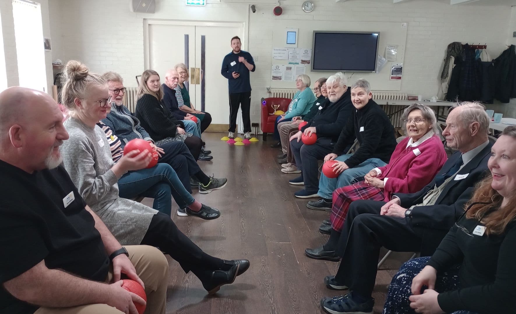 A group of older adults sit in two rows indoors, smiling and holding red balls, with an instructor standing. - Home Instead