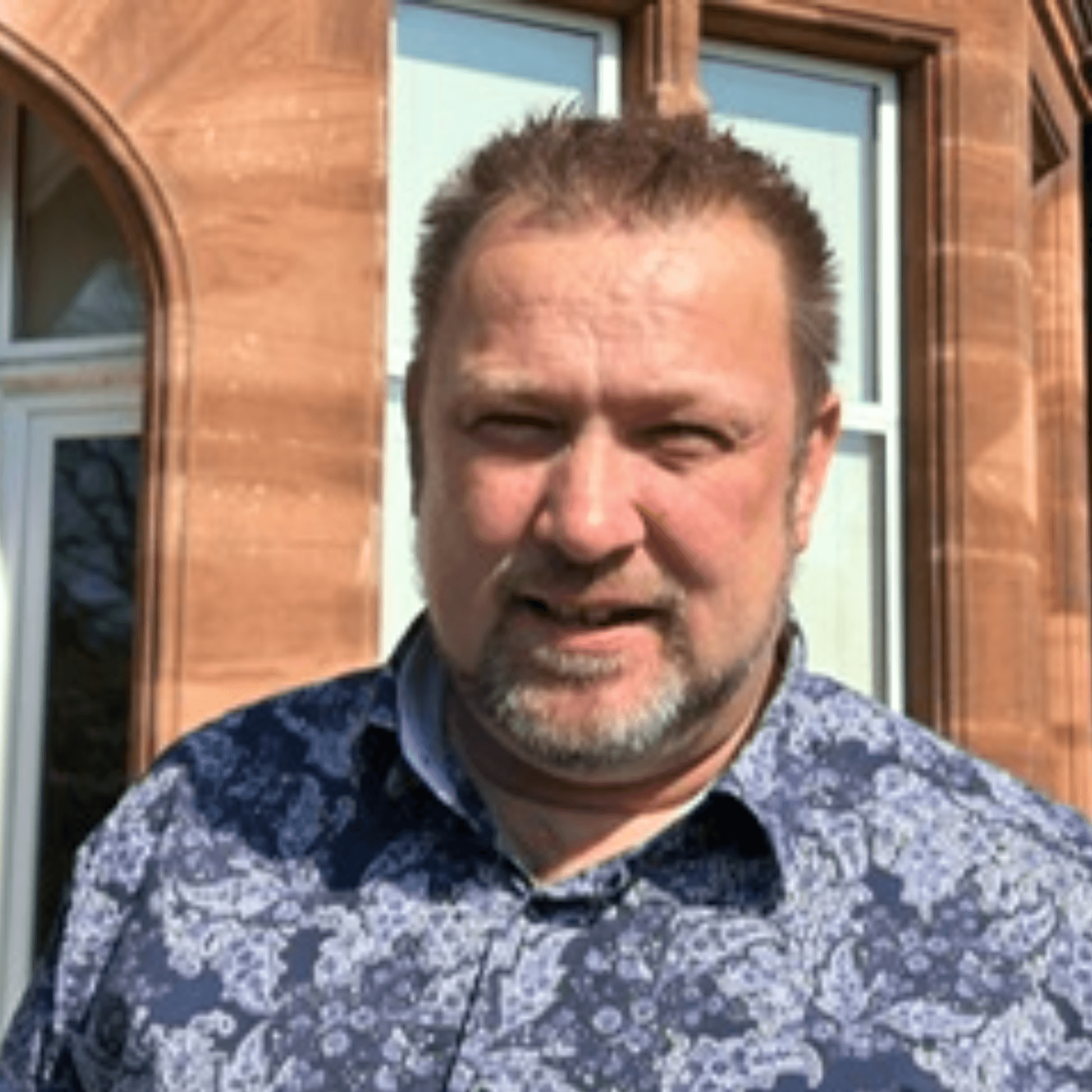 Man with short hair and beard wearing a blue patterned shirt stands outside in sunlight near a brick building. - Home Instead