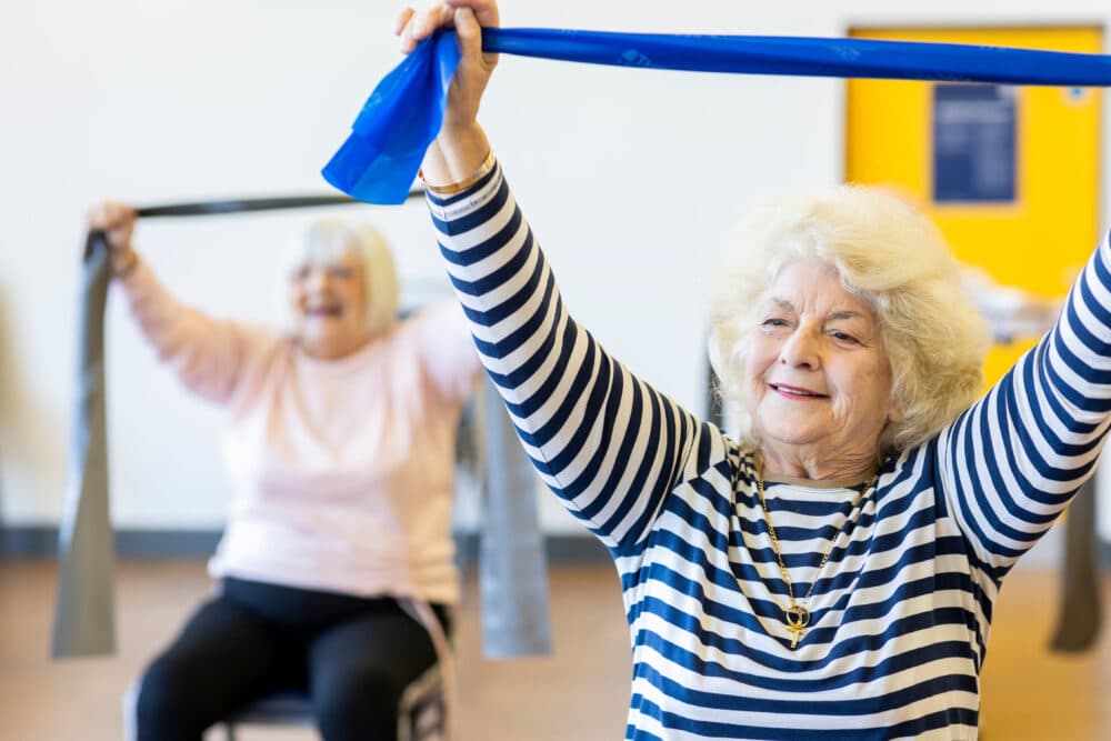 Two older women exercise with resistance bands in a bright room, smiling and raising their arms. - Home Instead