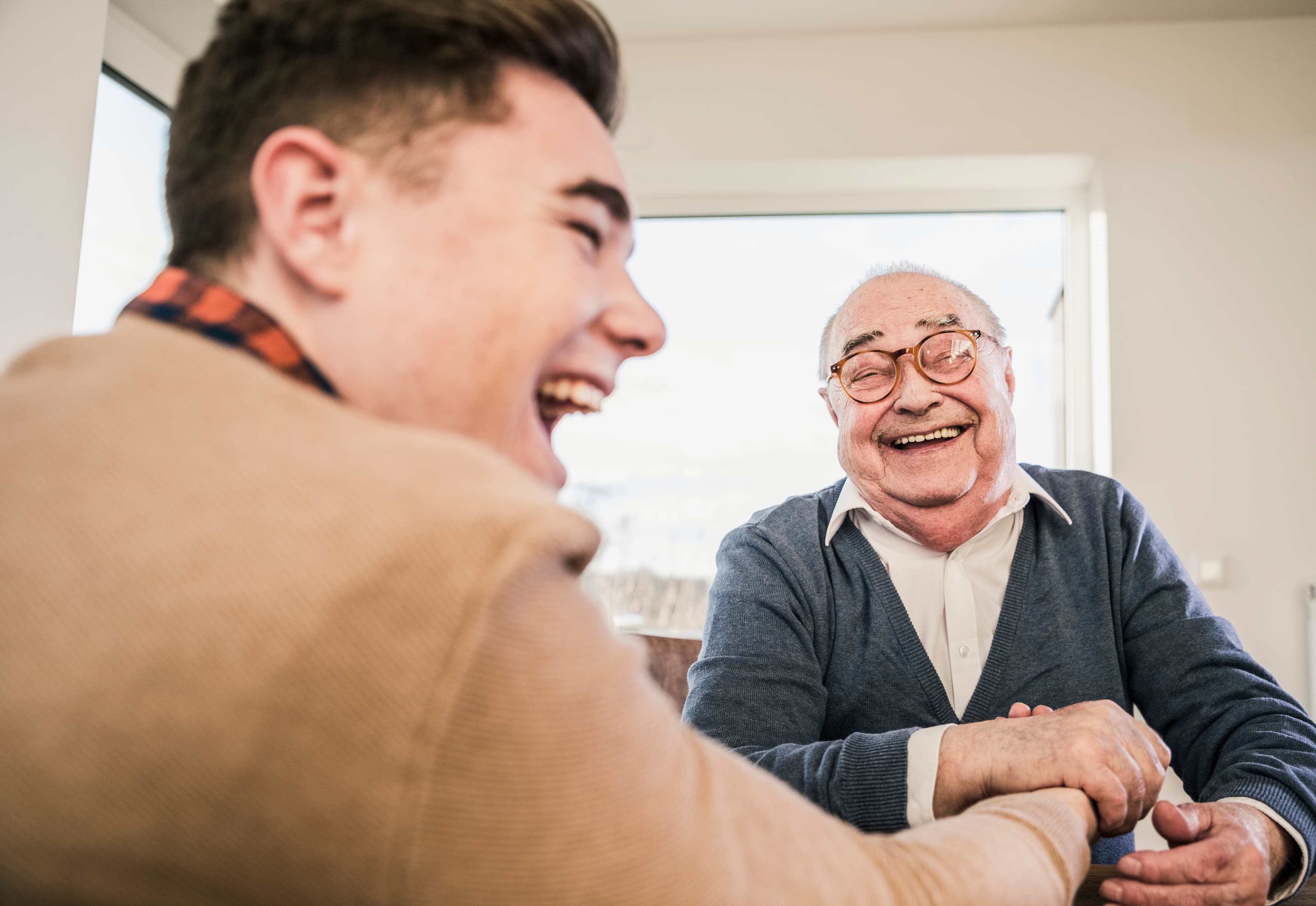 A young man and an elderly man laughing together at a table in a bright, cozy room. - Home Instead