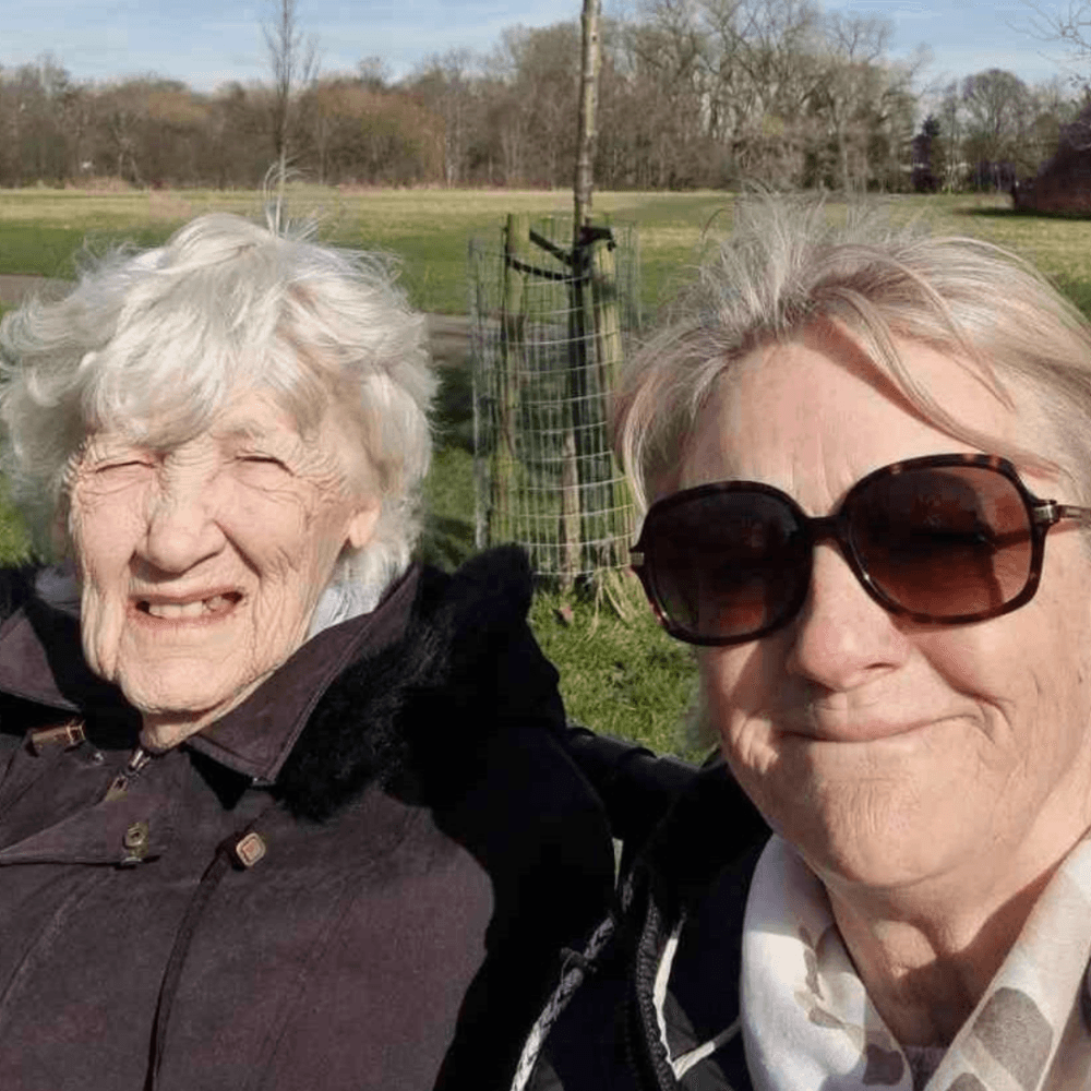 Two older women smiling outdoors in sunlight, with green grass and trees in the background. - Home Instead