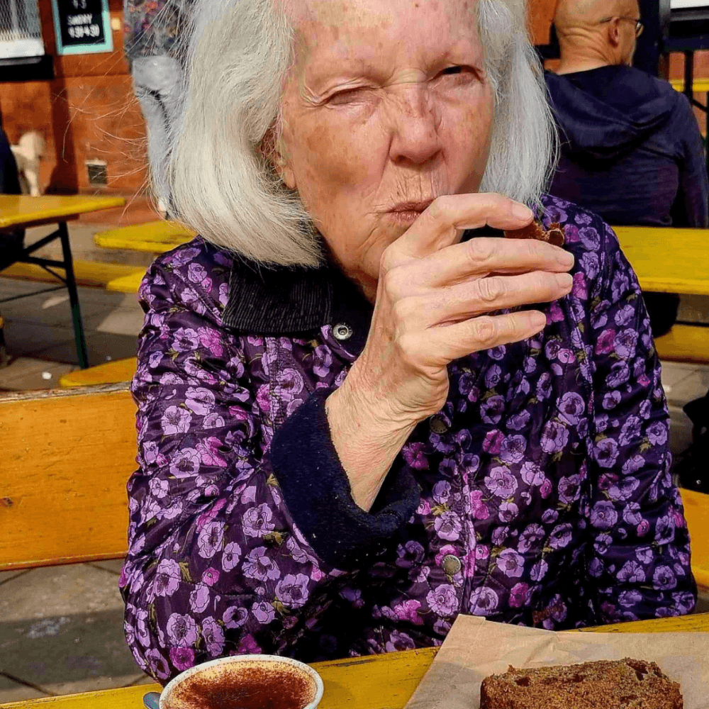 Older woman in a purple floral jacket eating cake and drinking coffee at an outdoor table. - Home Instead