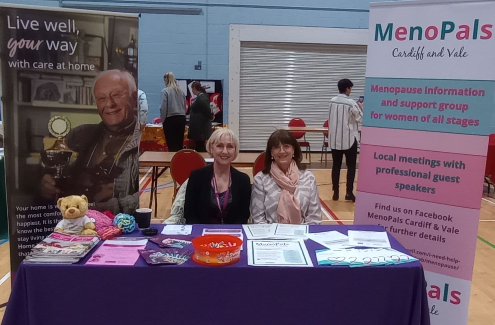 Two women sit at a Menopause info booth with banners, leaflets, and a teddy bear on a purple table. - Home Instead