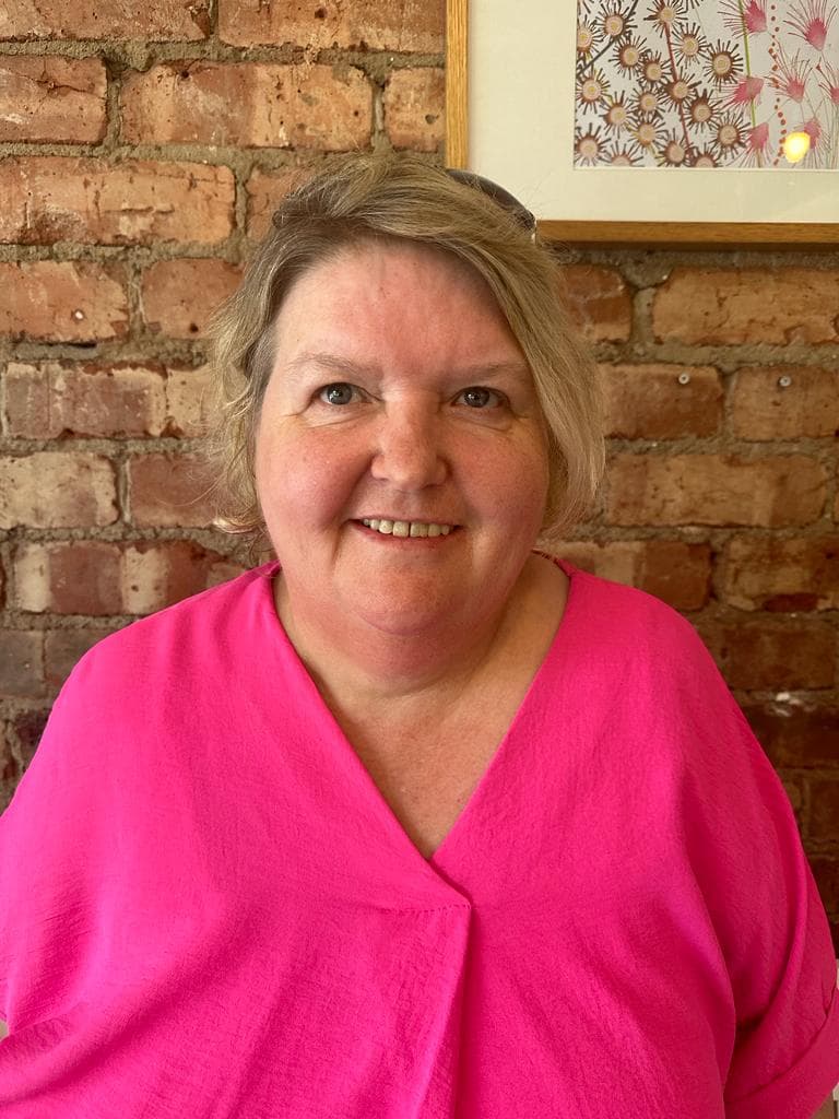 A smiling woman in a bright pink top stands in front of a brick wall and a framed floral picture. - Home Instead