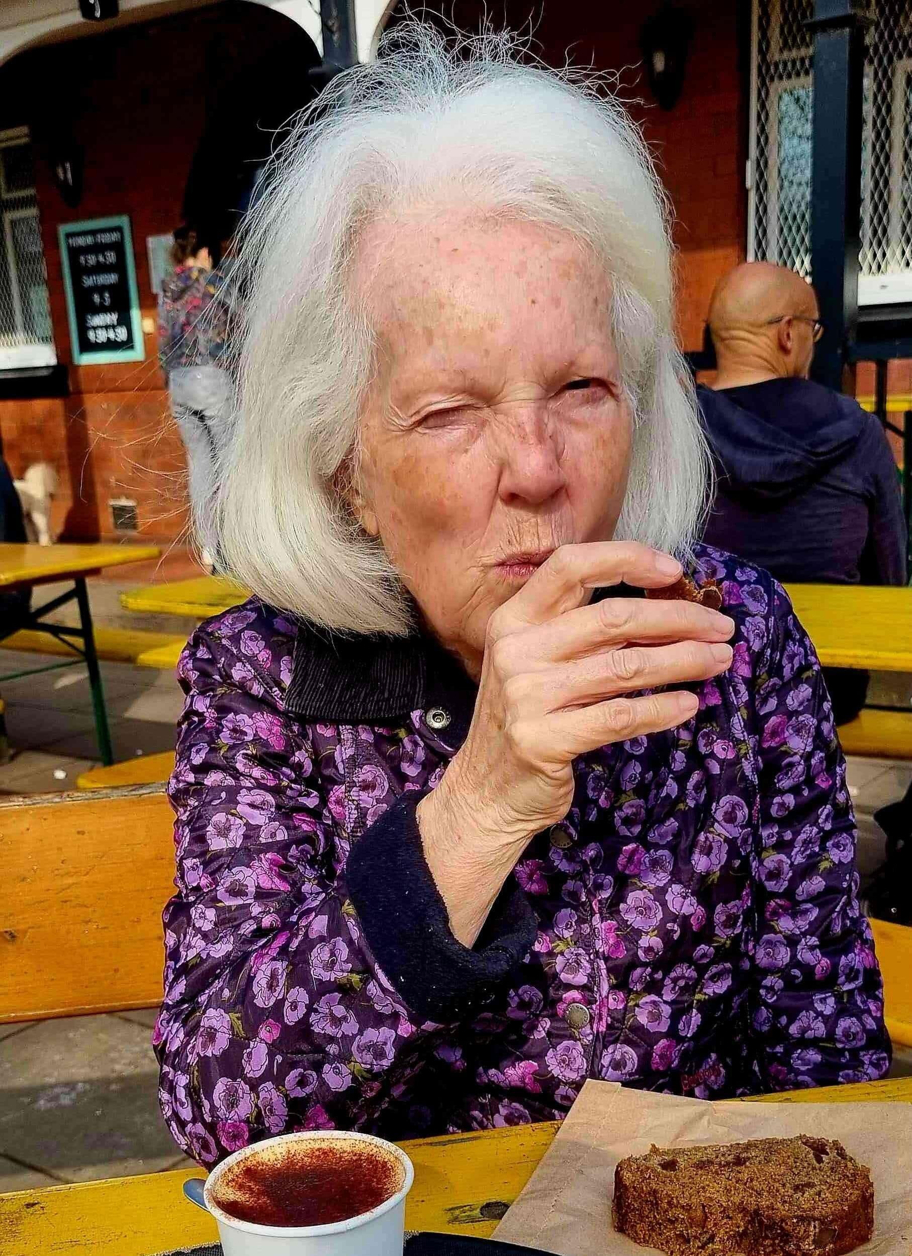 Older woman with white hair eating cake and drinking coffee at an outdoor cafe, wearing a purple floral jacket. - Home Instead