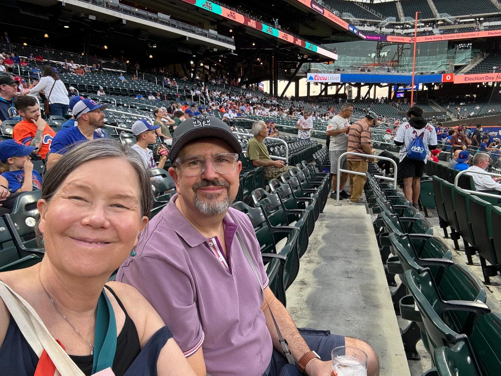 Two people smiling and sitting in stadium seats at a baseball game, with fans and the field in the background. - Home Instead