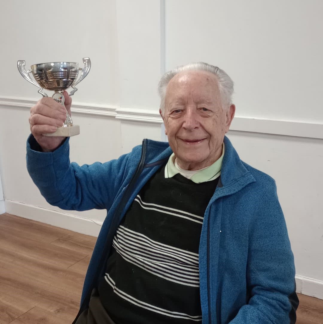Elderly man smiling and holding up a trophy while sitting indoors. - Home Instead