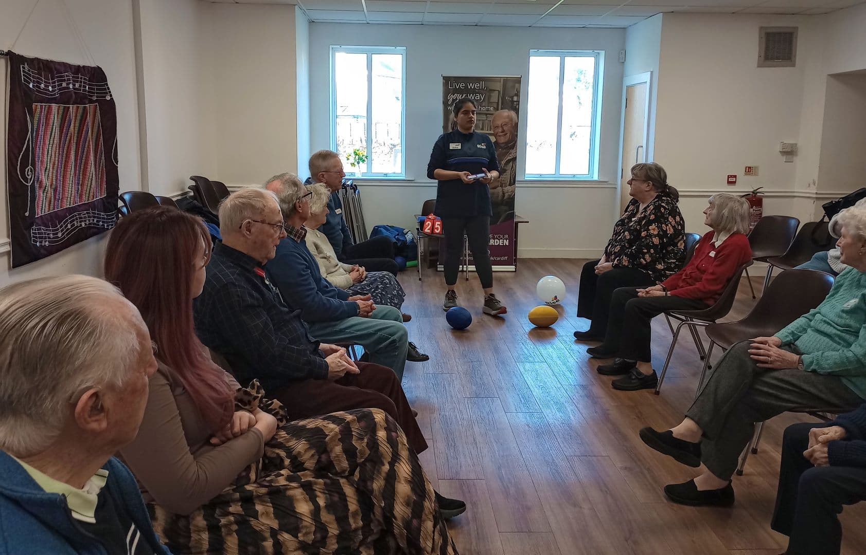 Elderly people sit in a circle indoors, with two staff standing and balloons in the center of the room. - Home Instead
