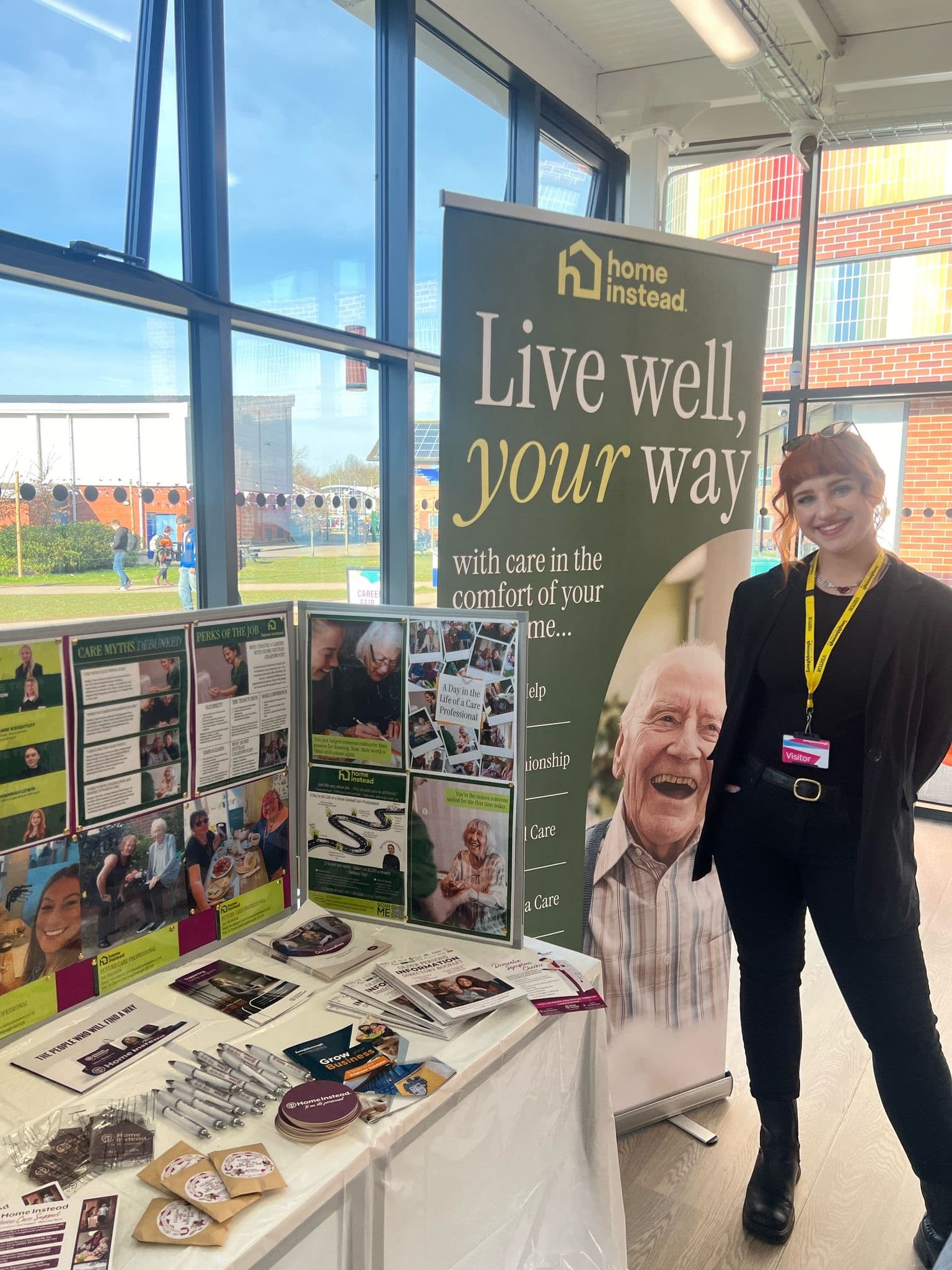 A woman stands by a display table for Home Instead, promoting in-home elderly care at an indoor event. - Home Instead