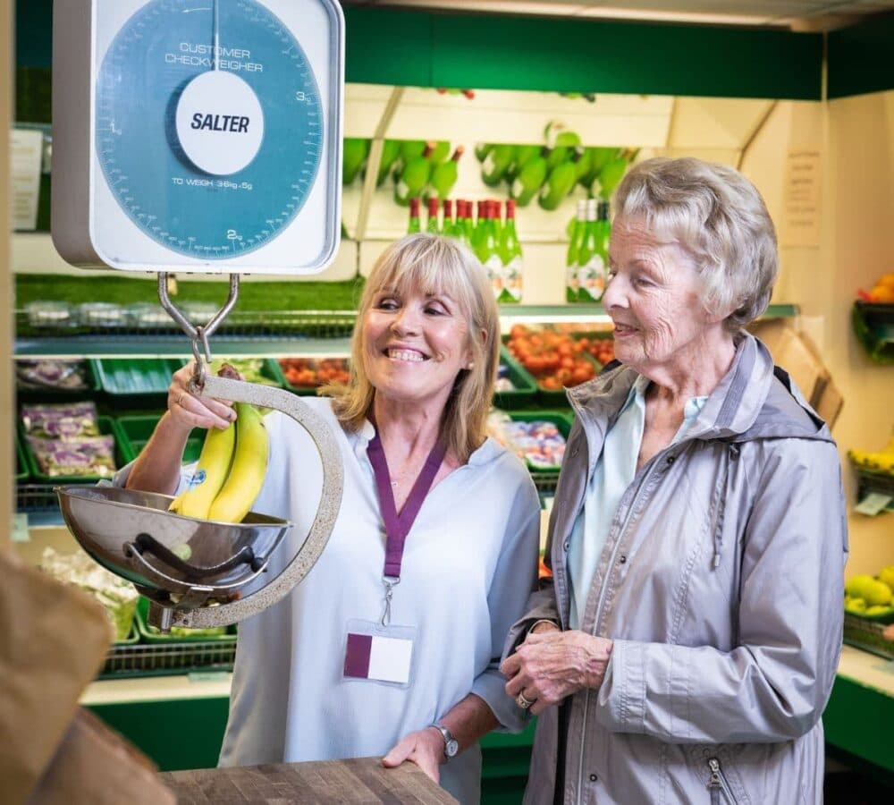 A younger female carer with short blonde hair weighing a banana with an older female adult with short hair wearing sweater inside the grocery