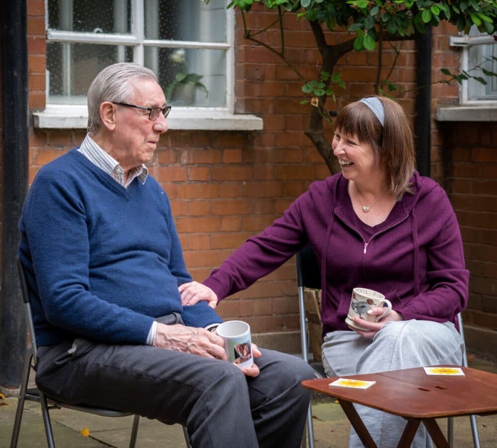 An older man with grey hair, wearing eyeglasses and blue tops with his younger carer with short hair and purple sweater bot sitting outside the house and happy and smiling