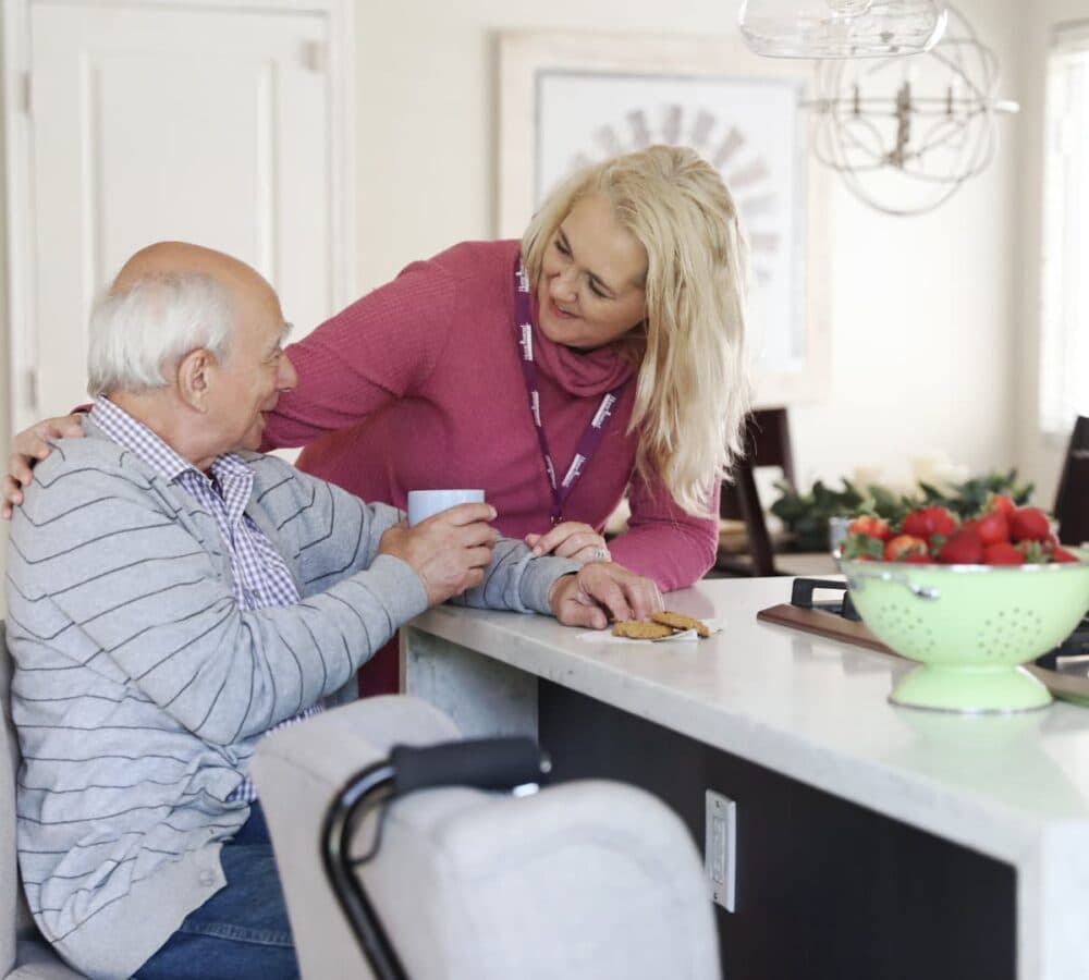 Older male adult inside the kitchen with white hair while drinking water with his younger female carer with long hair and wearing pink both happy and smiling