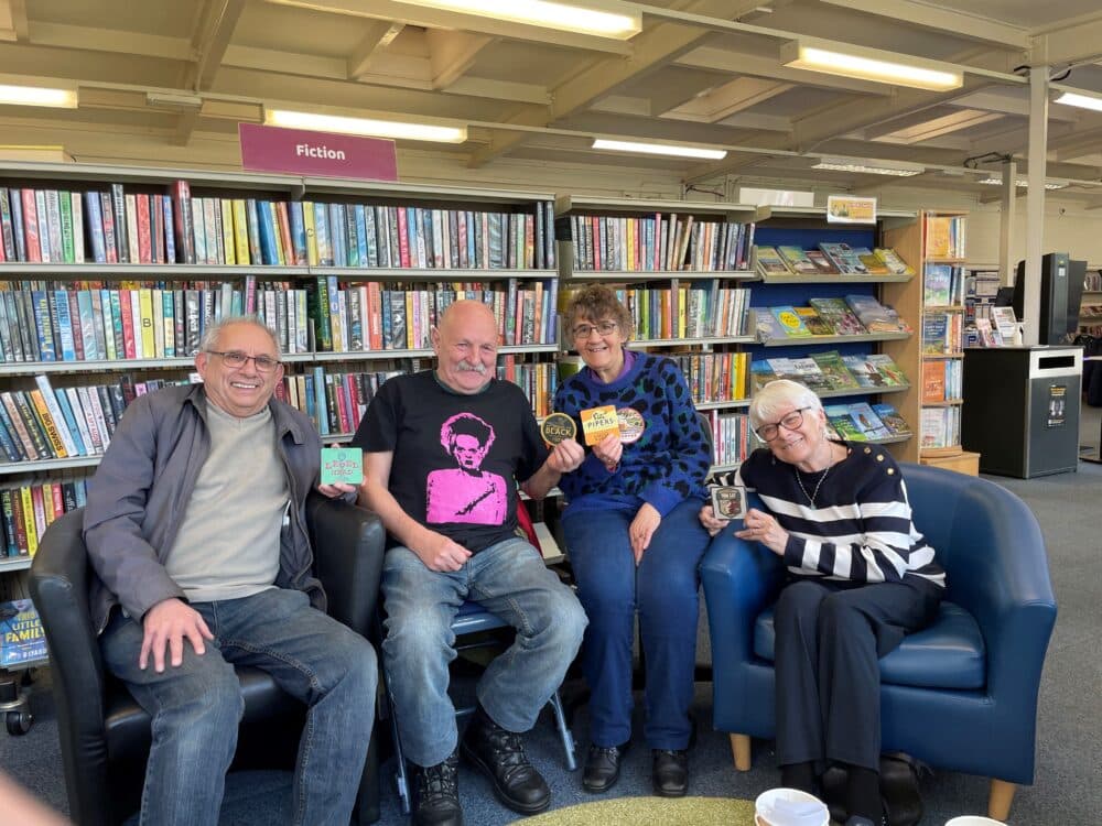 Four smiling adults sit in a library, each holding a book, with shelves of books in the background. - Home Instead