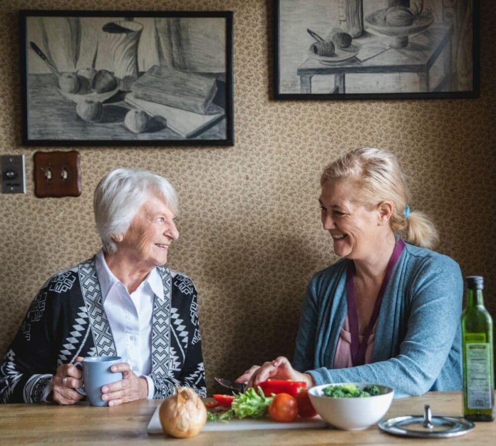 An older woman holding a cup of coffee while chatting with her younger female carer while chopping vegetables inside the kitchen
