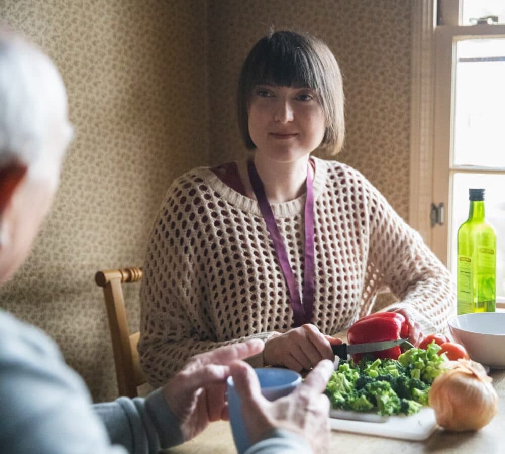 Woman chopping vegetables at a kitchen table, talking with another person holding a cup. - Home Instead