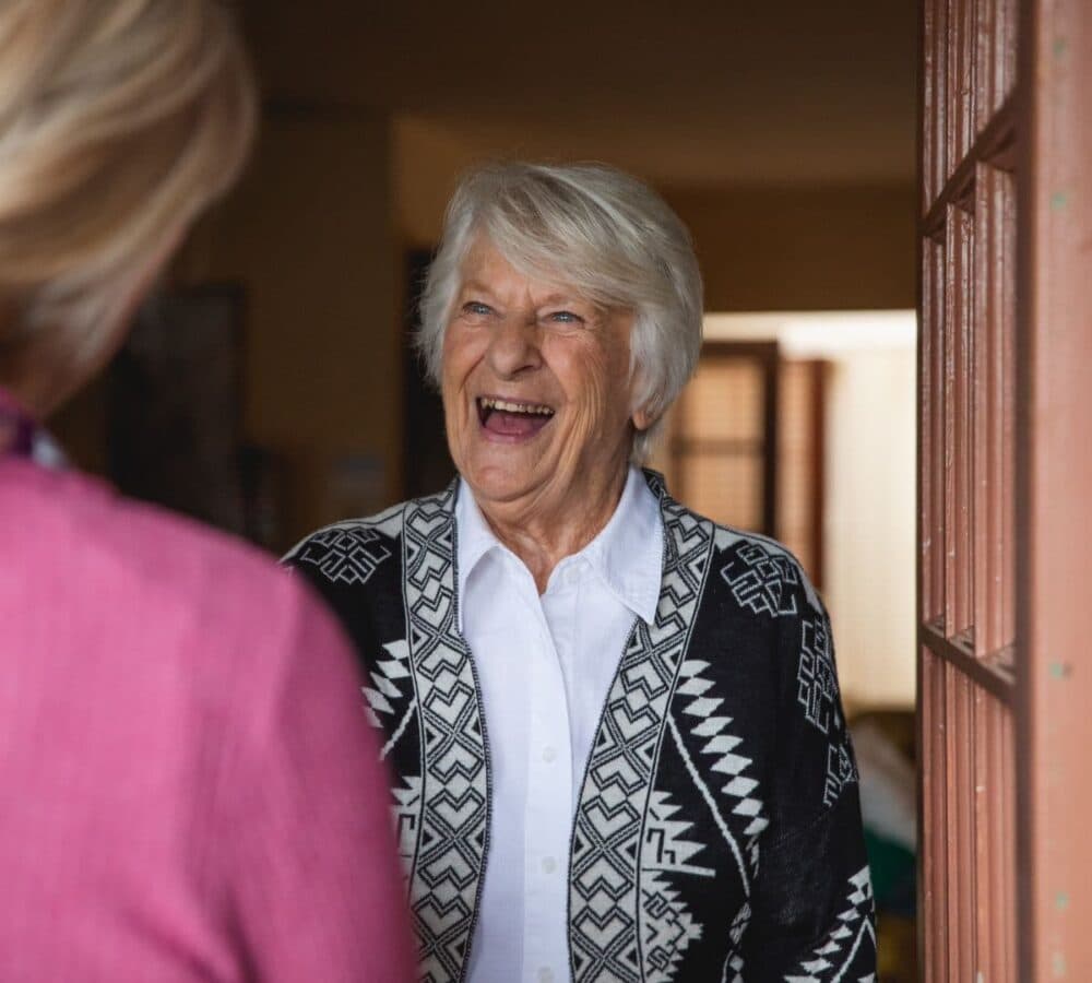 Older woman wearing short grey hair happy and has a big smile while opening the door for her Care Professional from Home Instead