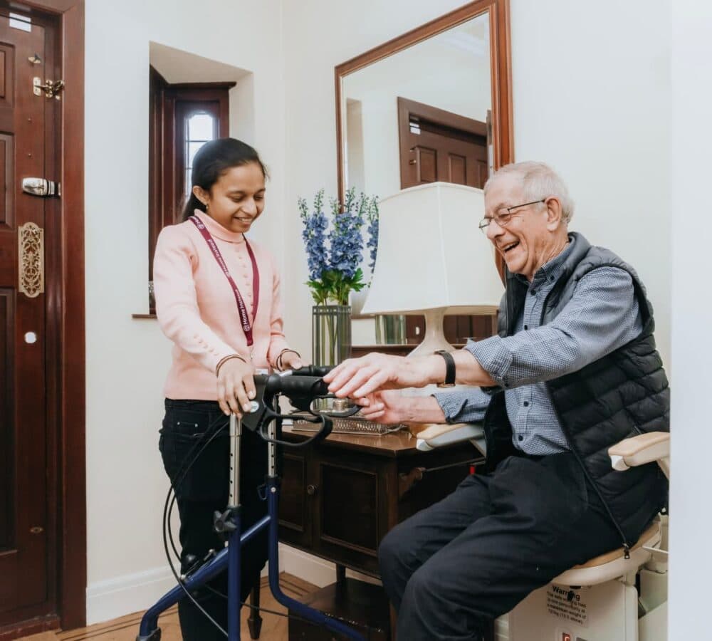 A male older adult trying to use a walker with the help of his younger female carer inside his home